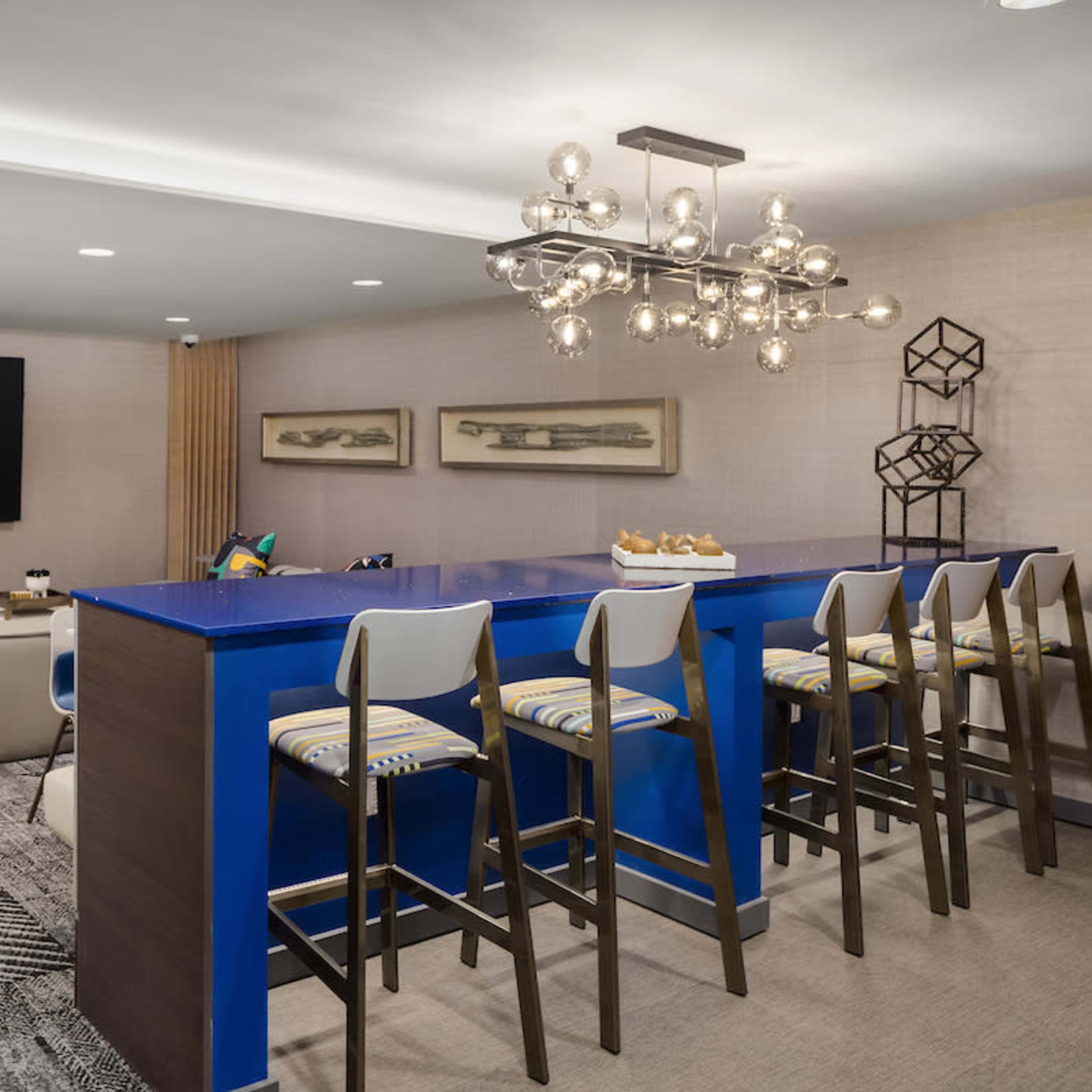 A modern lounge area featuring a blue countertop bar with four high stools, a large television on the wall, and a seating arrangement with a light-colored couch.