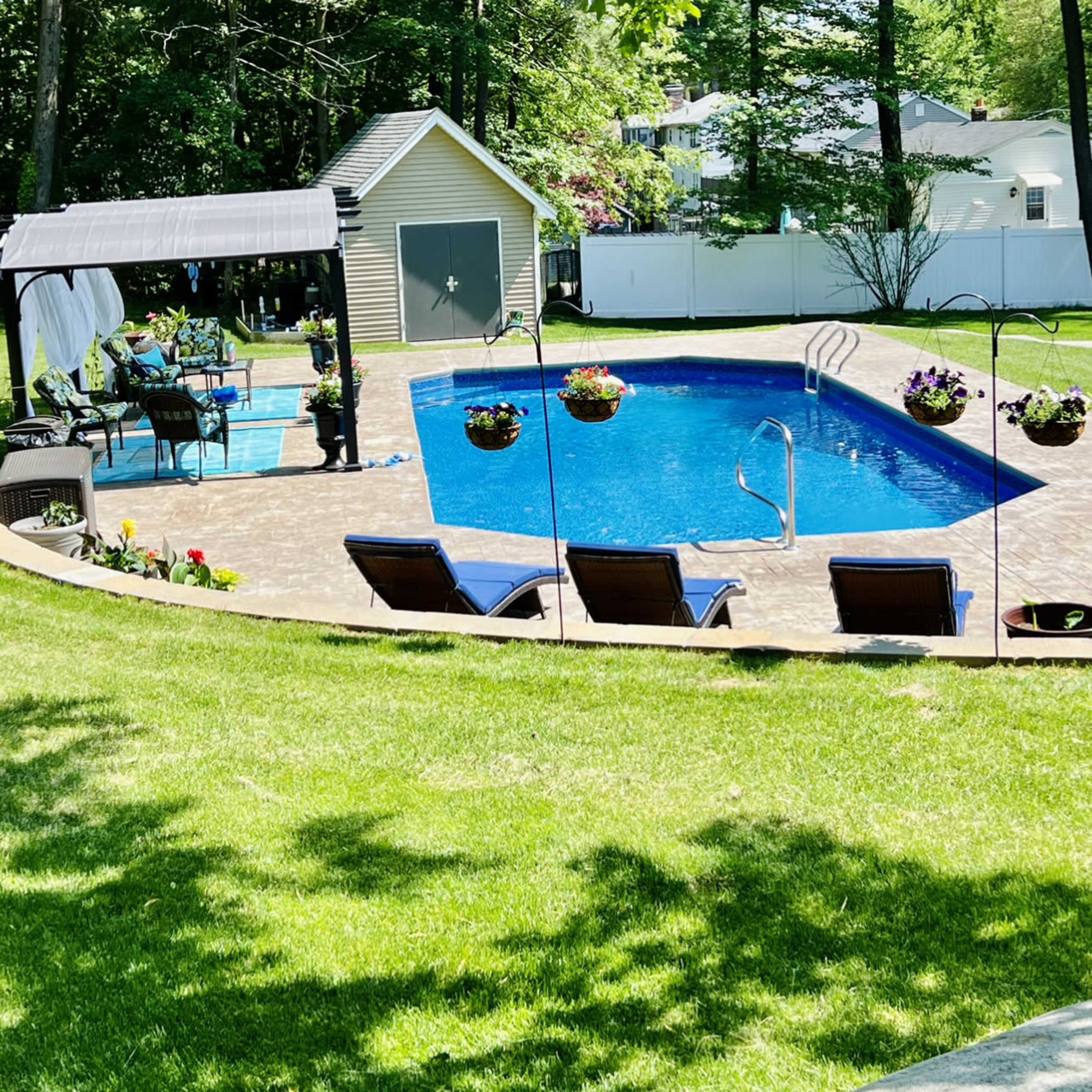 A backyard with a blue swimming pool, lounge chairs, a gazebo, and a small shed, surrounded by trees and a white fence.