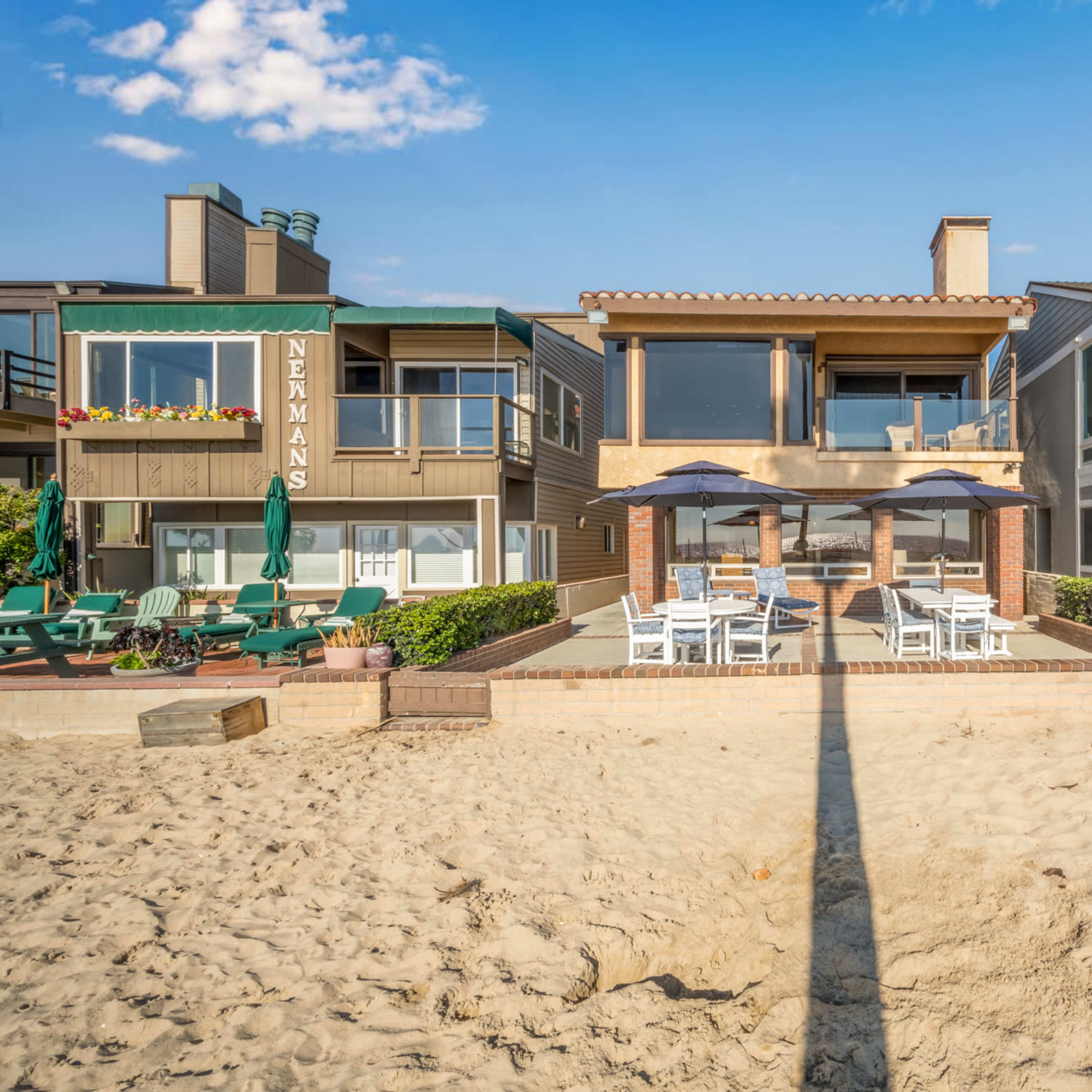 A beachfront property with two houses flanking a sandy pathway leading to a terrace with seating under a shaded awning.
