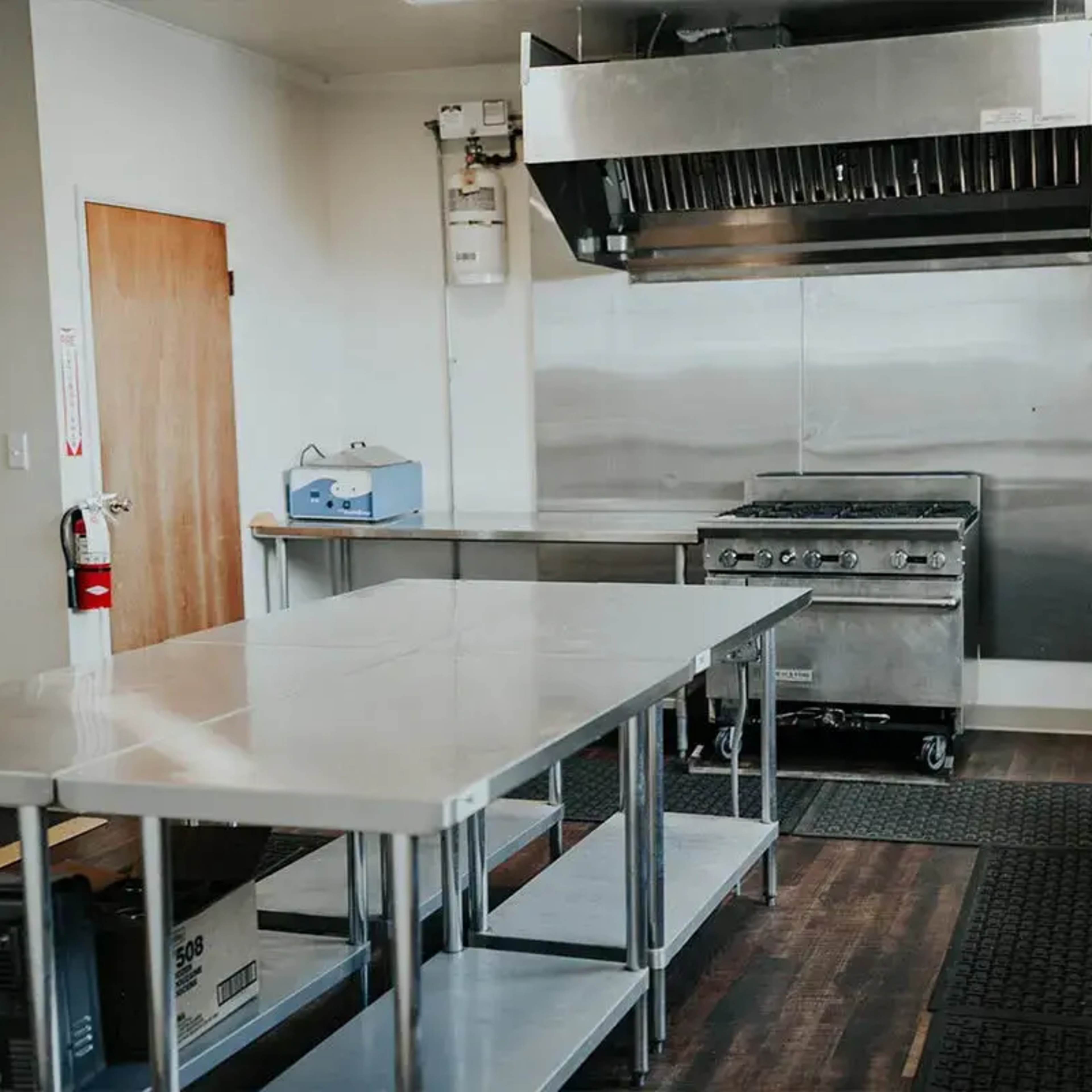A commercial kitchen with stainless steel appliances, including a stove and exhaust hood, along with several stainless steel tables and rubber mats on the floor.