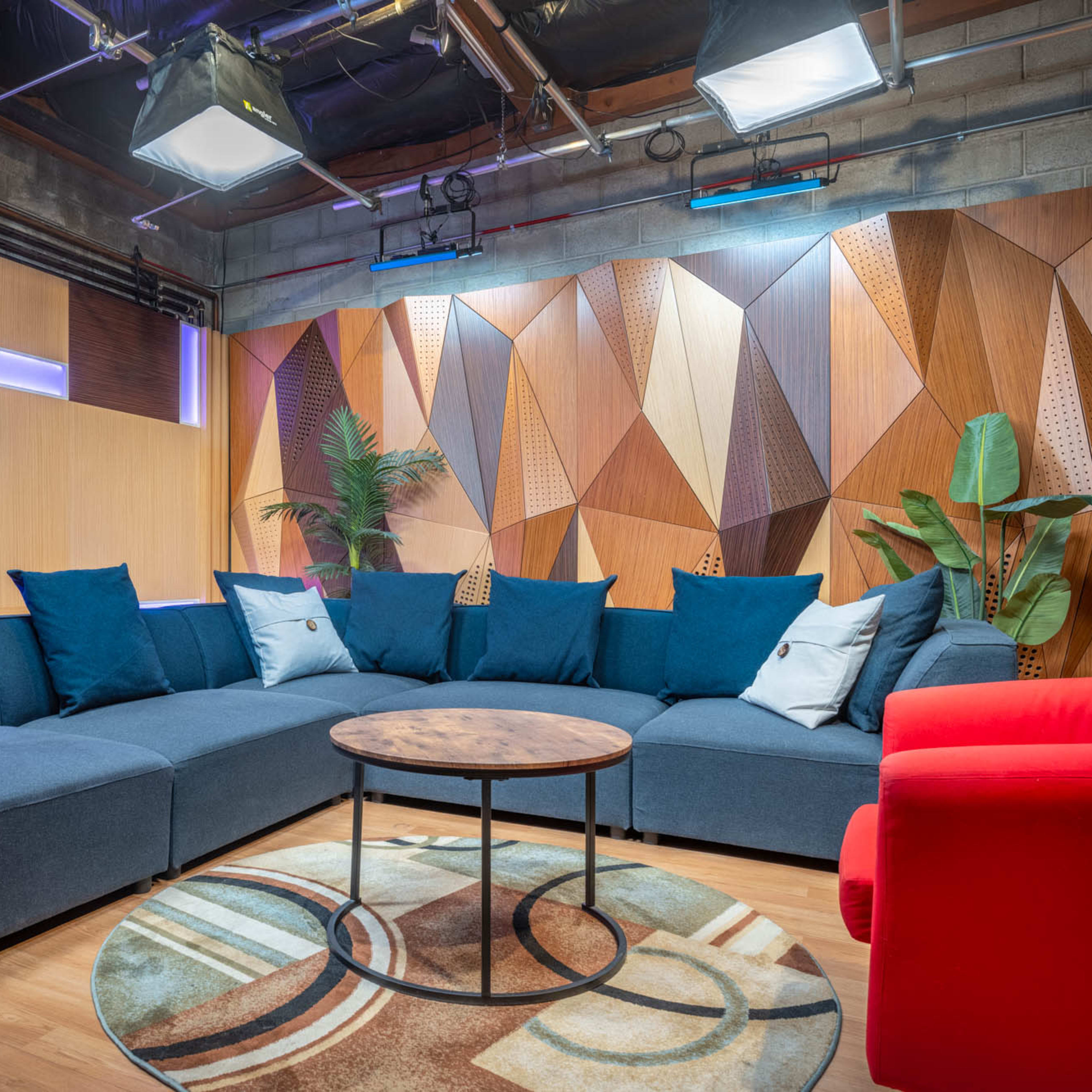 A modern lounge area featuring a large sectional sofa with blue cushions, a round wooden coffee table, and a red accent chair, all set against a geometric wall design and indoor plants.