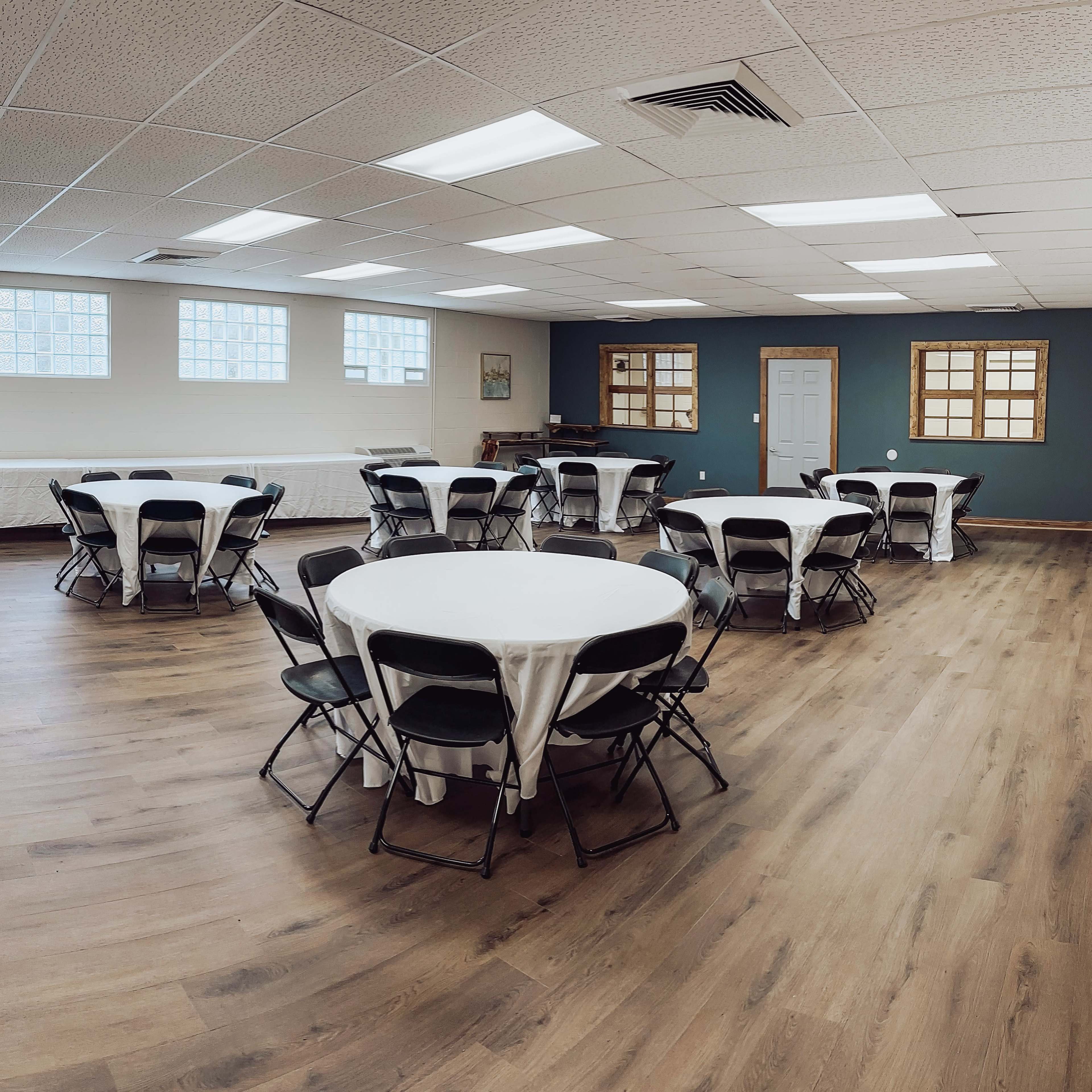 A spacious indoor venue features several round tables with white tablecloths and black chairs arranged on a polished wooden floor.
