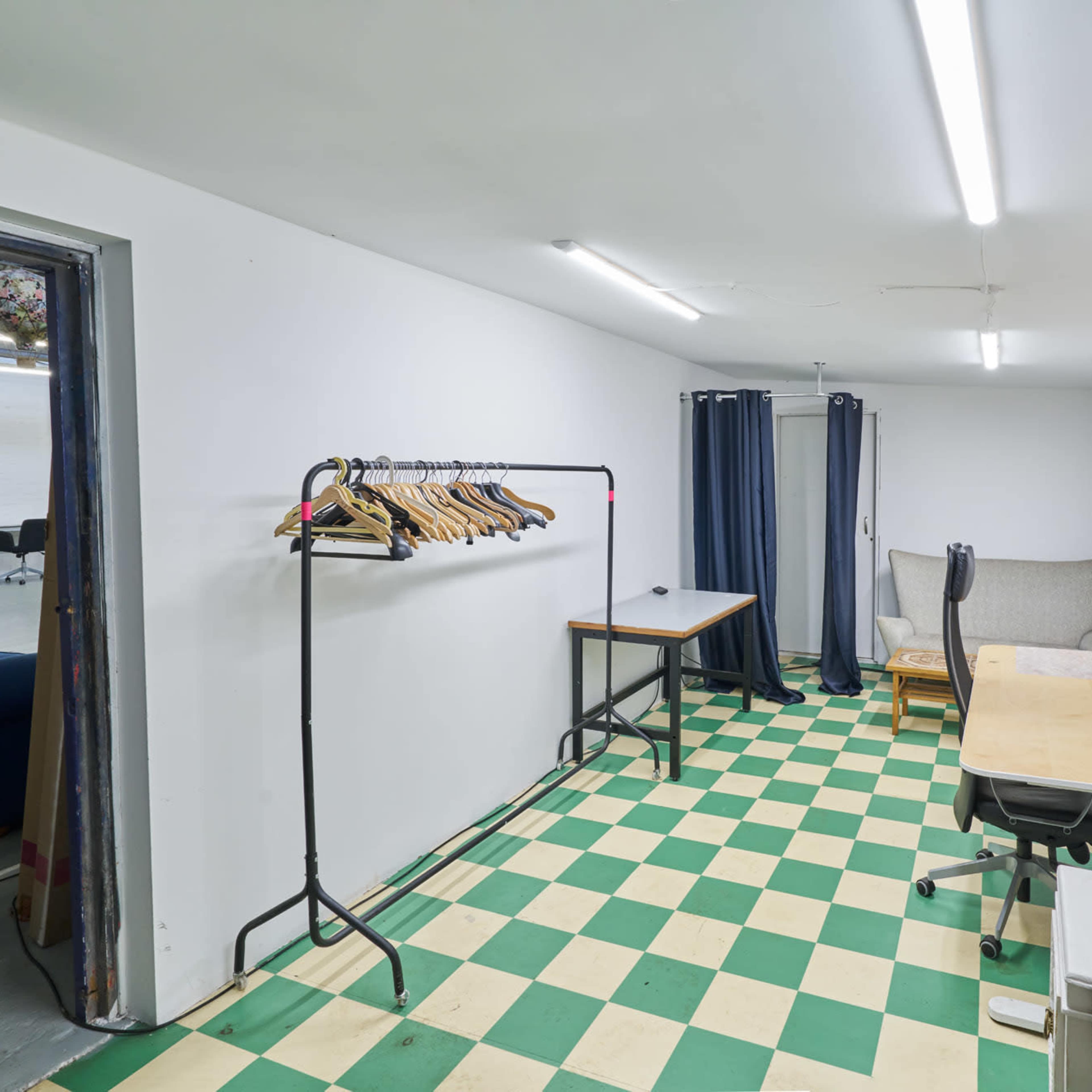 A small room with green and white checkered flooring, featuring a clothing rack, a table, a chair, and gray curtains.