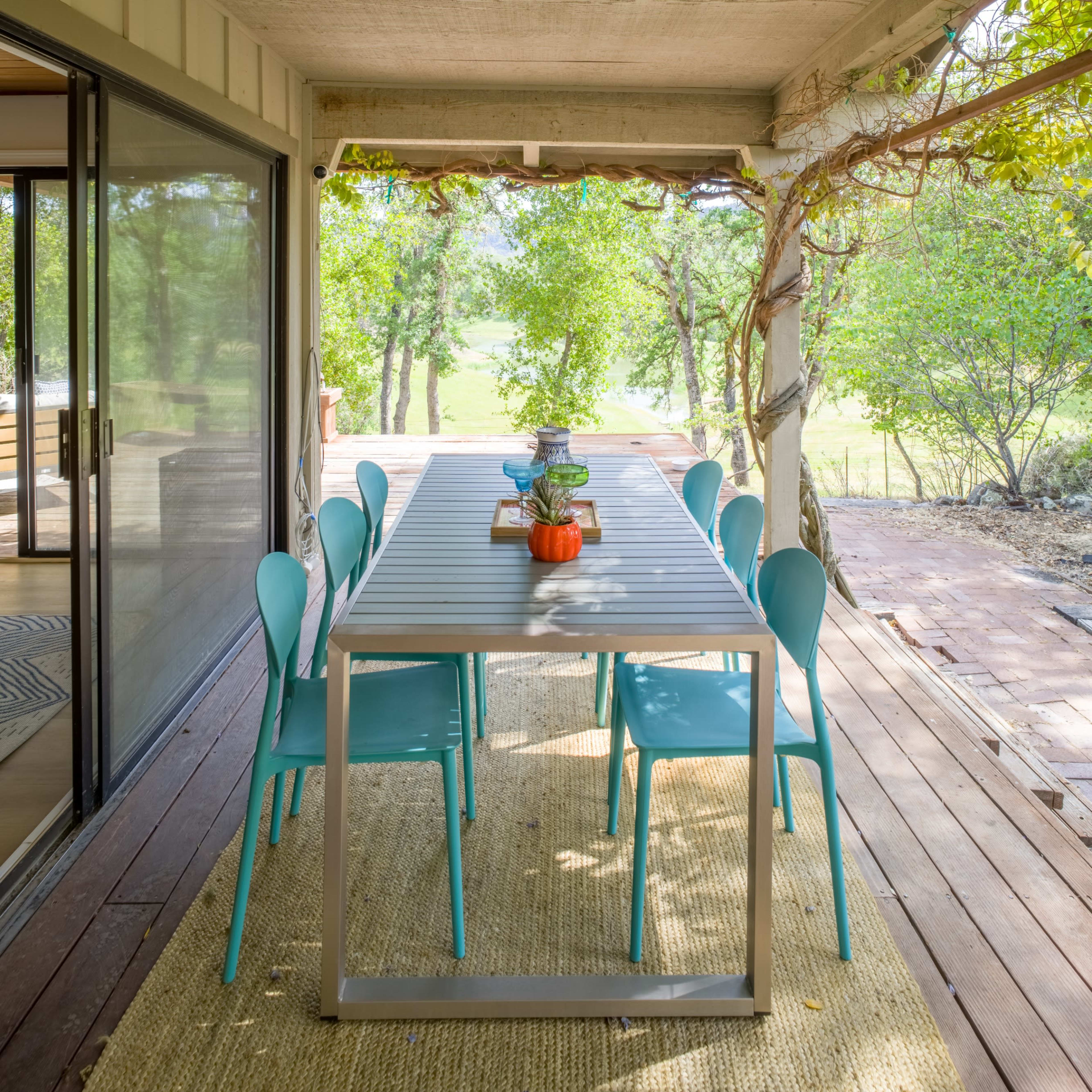 A long dining table with blue chairs is situated on a porch surrounded by greenery.