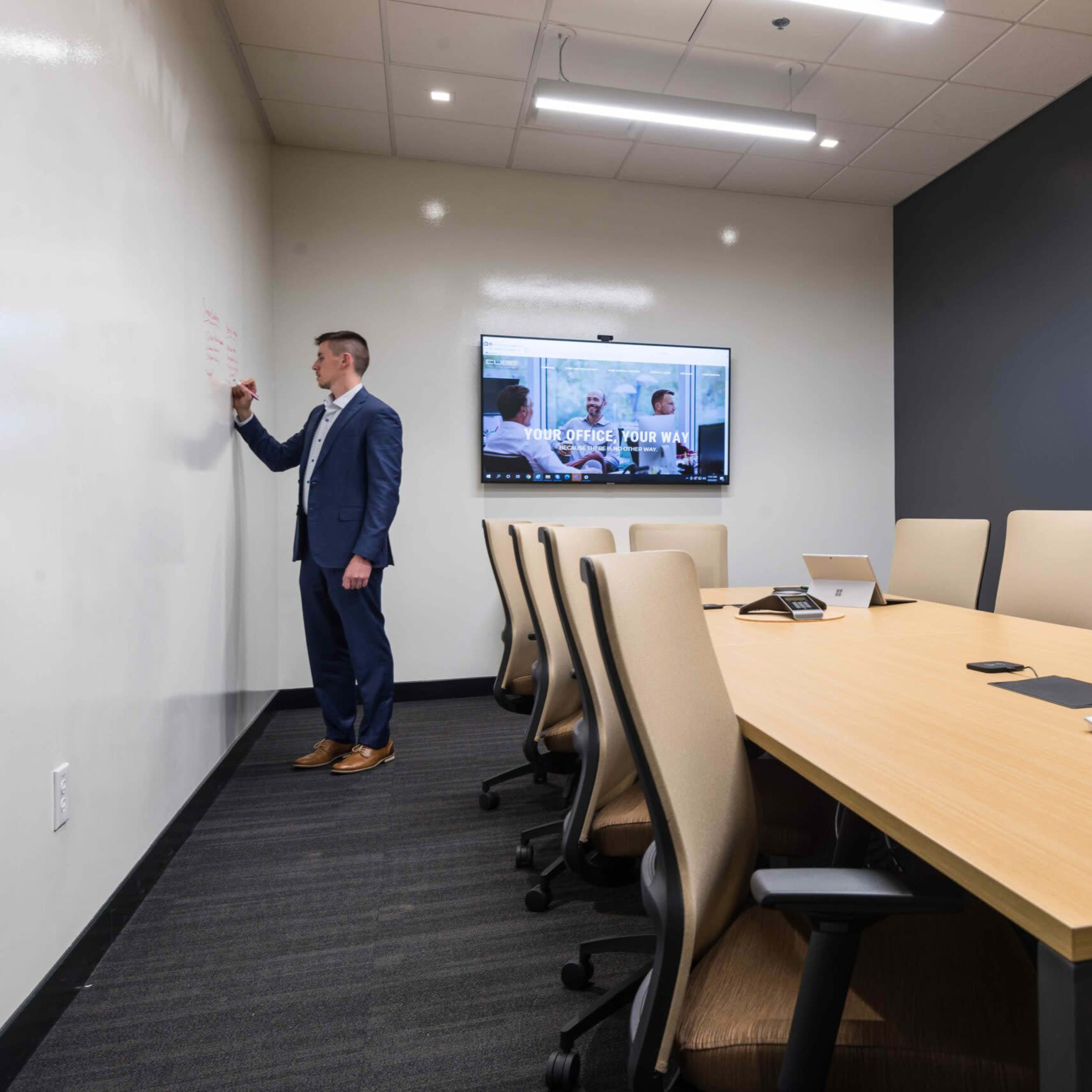 A man in formal attire writes on a whiteboard in a modern conference room equipped with a large table and a television screen.