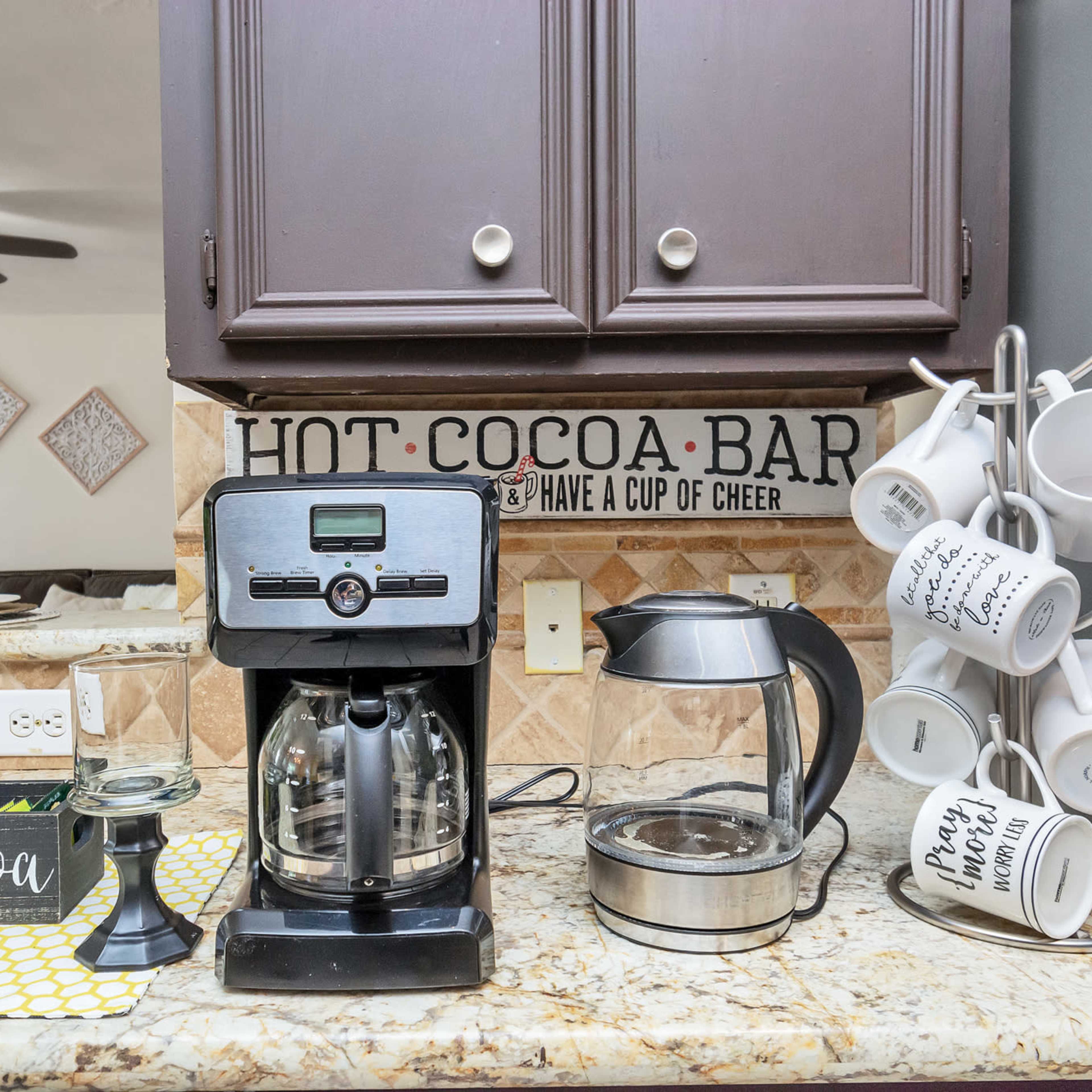 A kitchen countertop with a coffee machine, a kettle, and a mug holder next to a decorative sign that reads "HOT COCOA BAR."