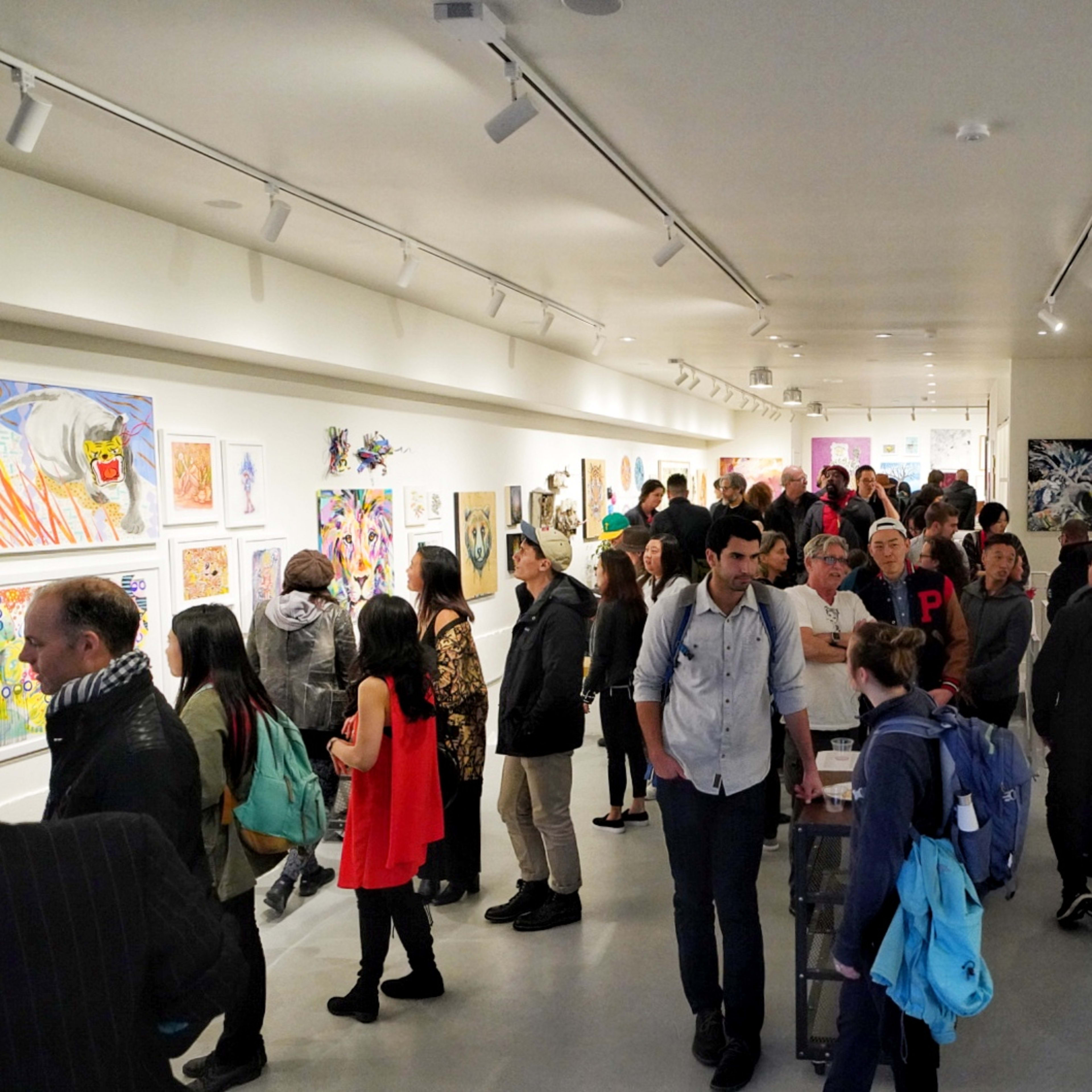 A crowded art gallery features people viewing various artworks displayed on the walls.