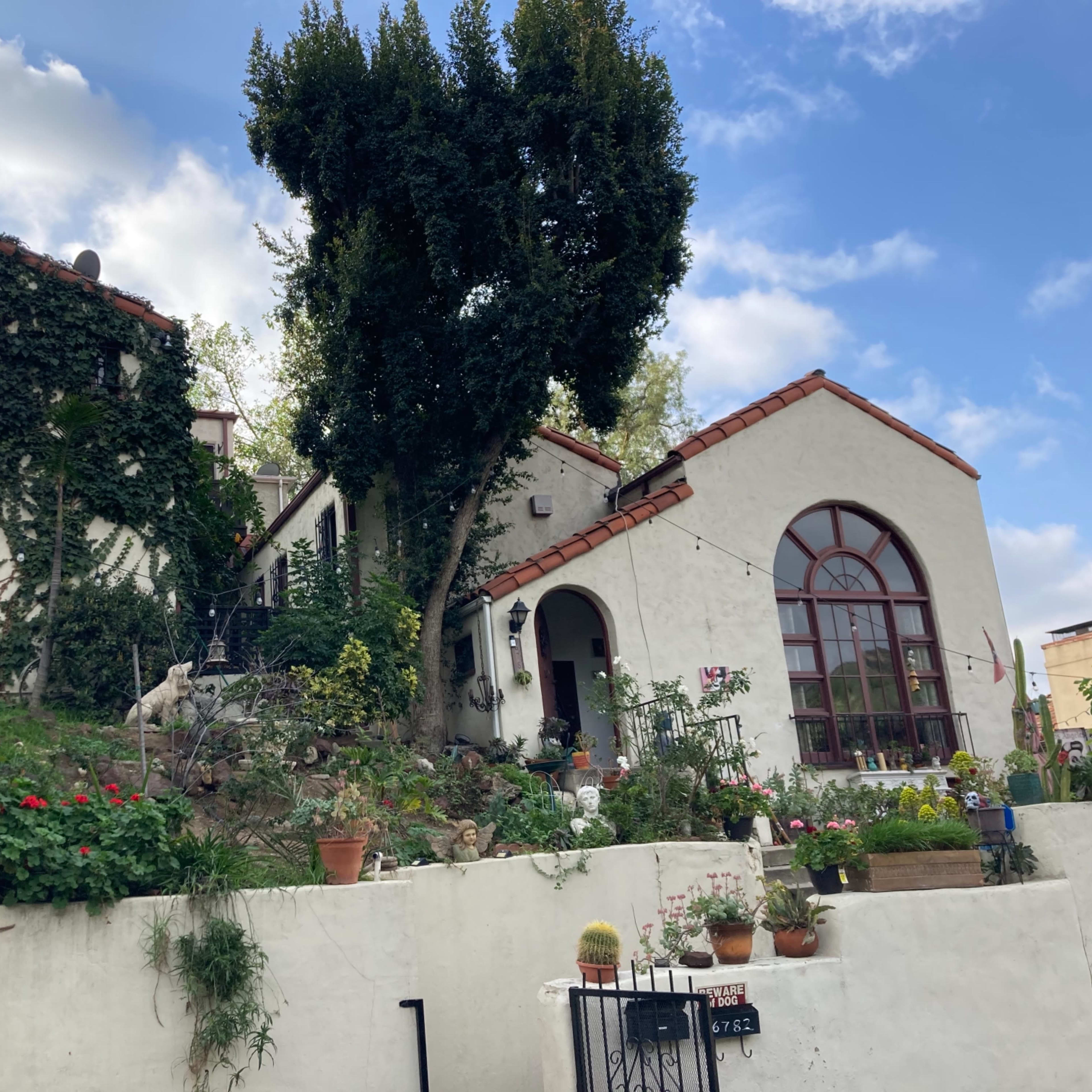 A hillside house with white walls, red-tiled roofs, and various plants in pots surrounding the entrance.