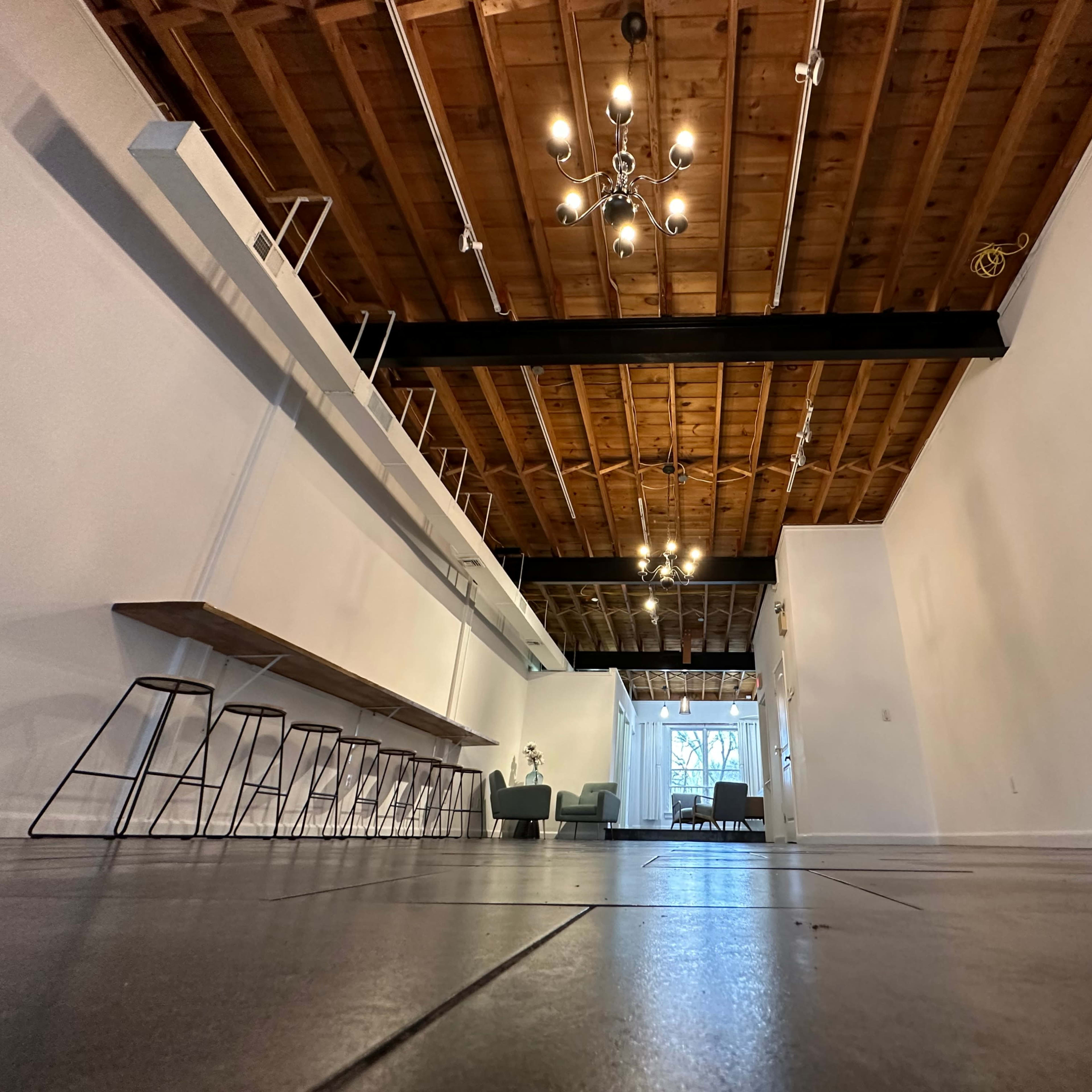 A spacious interior with exposed wooden beams, a long bar area with stools, and a wide entrance leading to a well-lit area.