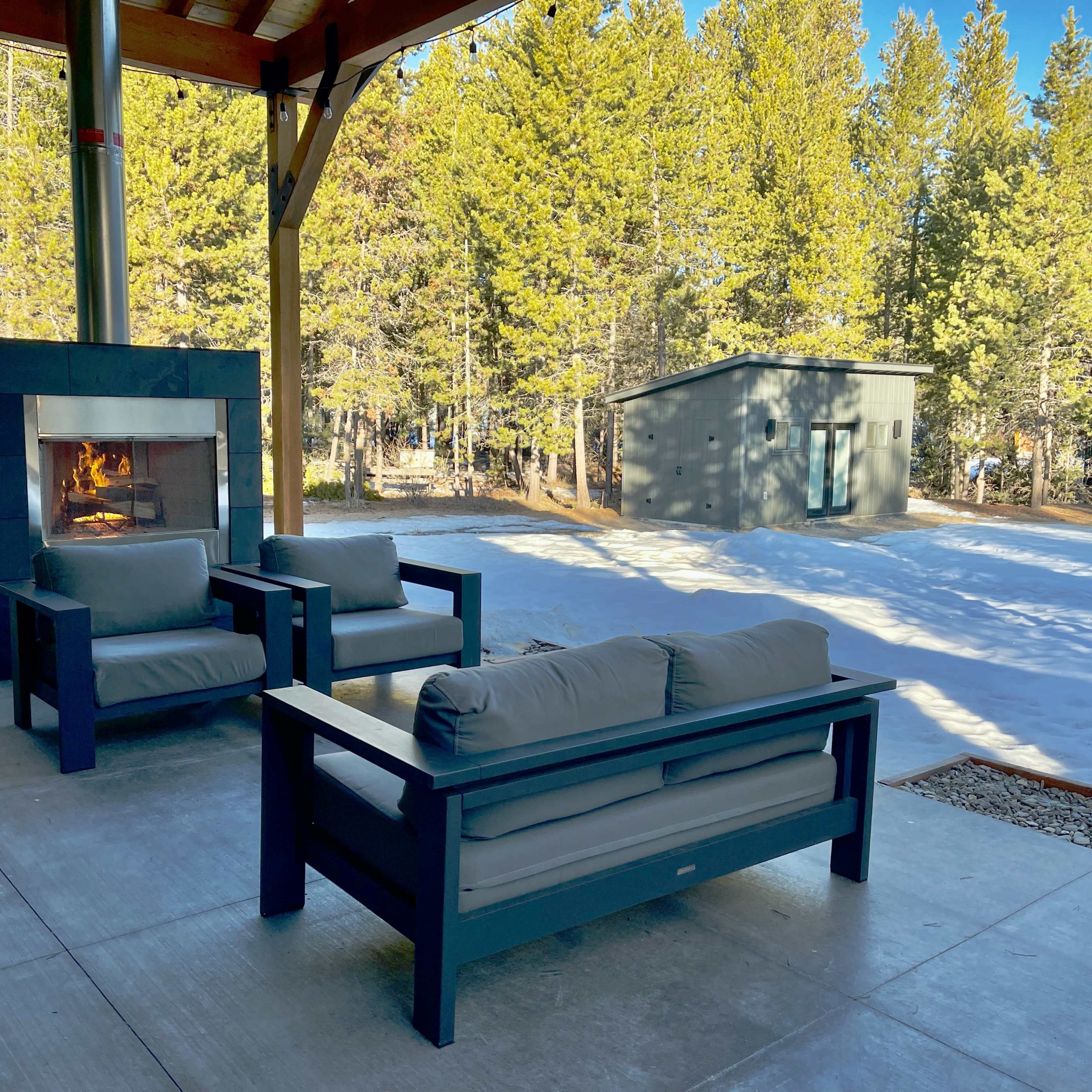 A seating area with couch and chairs is arranged beside a modern outdoor fireplace, surrounded by trees and a patch of snow.