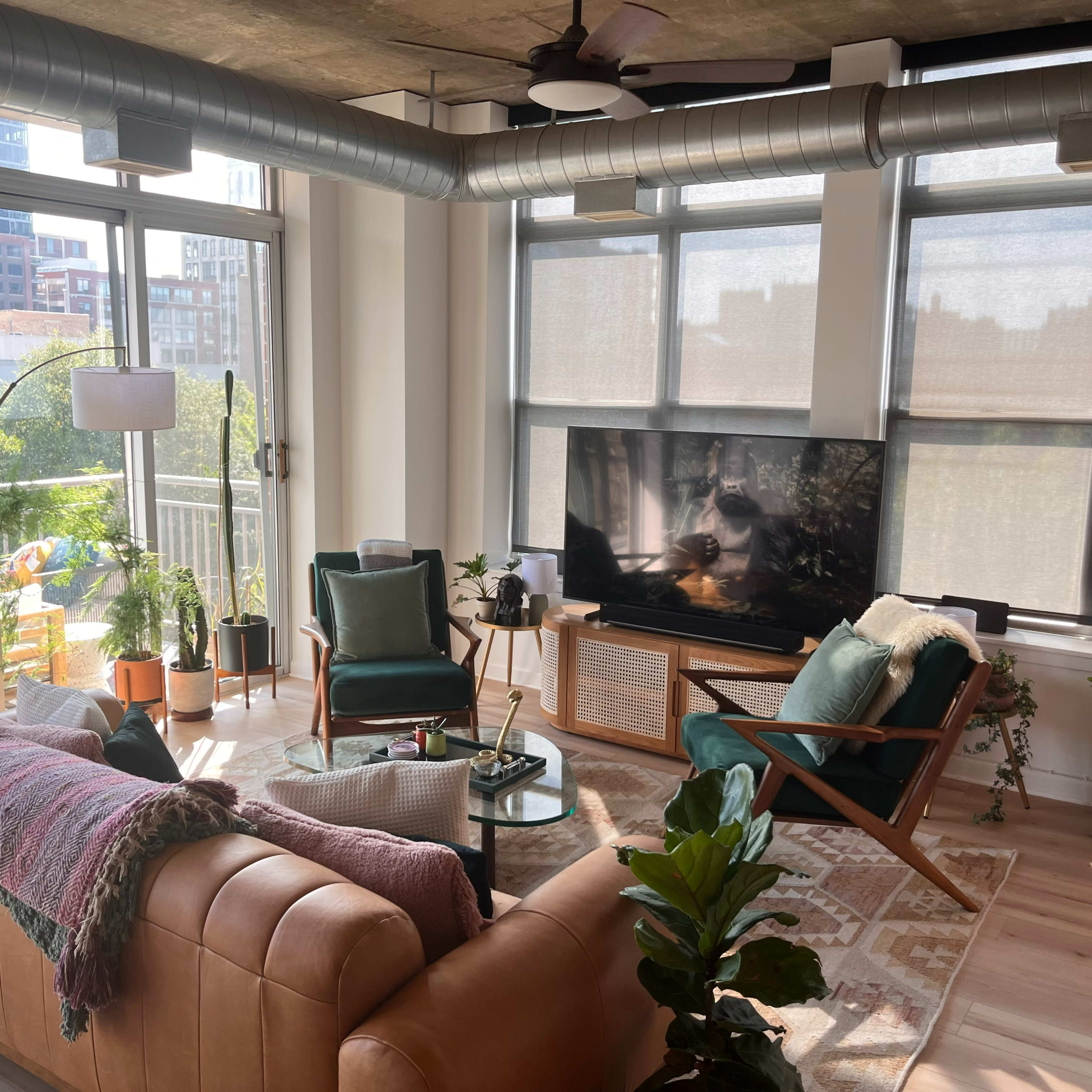 A modern living room with a large sofa, plants, a television on a stand, and large windows letting in natural light.