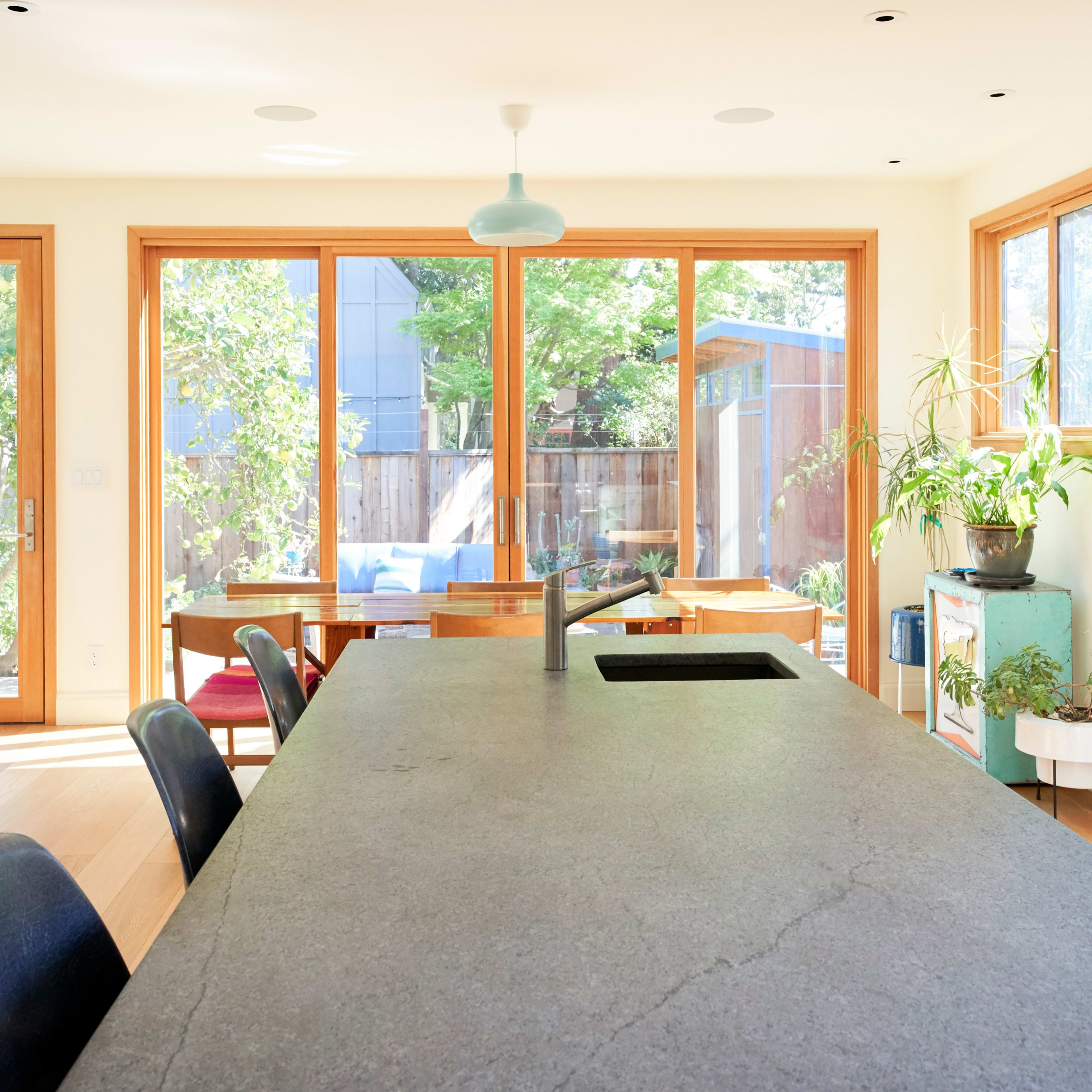 A modern kitchen with a large stone island, large windows overlooking a garden, and minimalist decor.