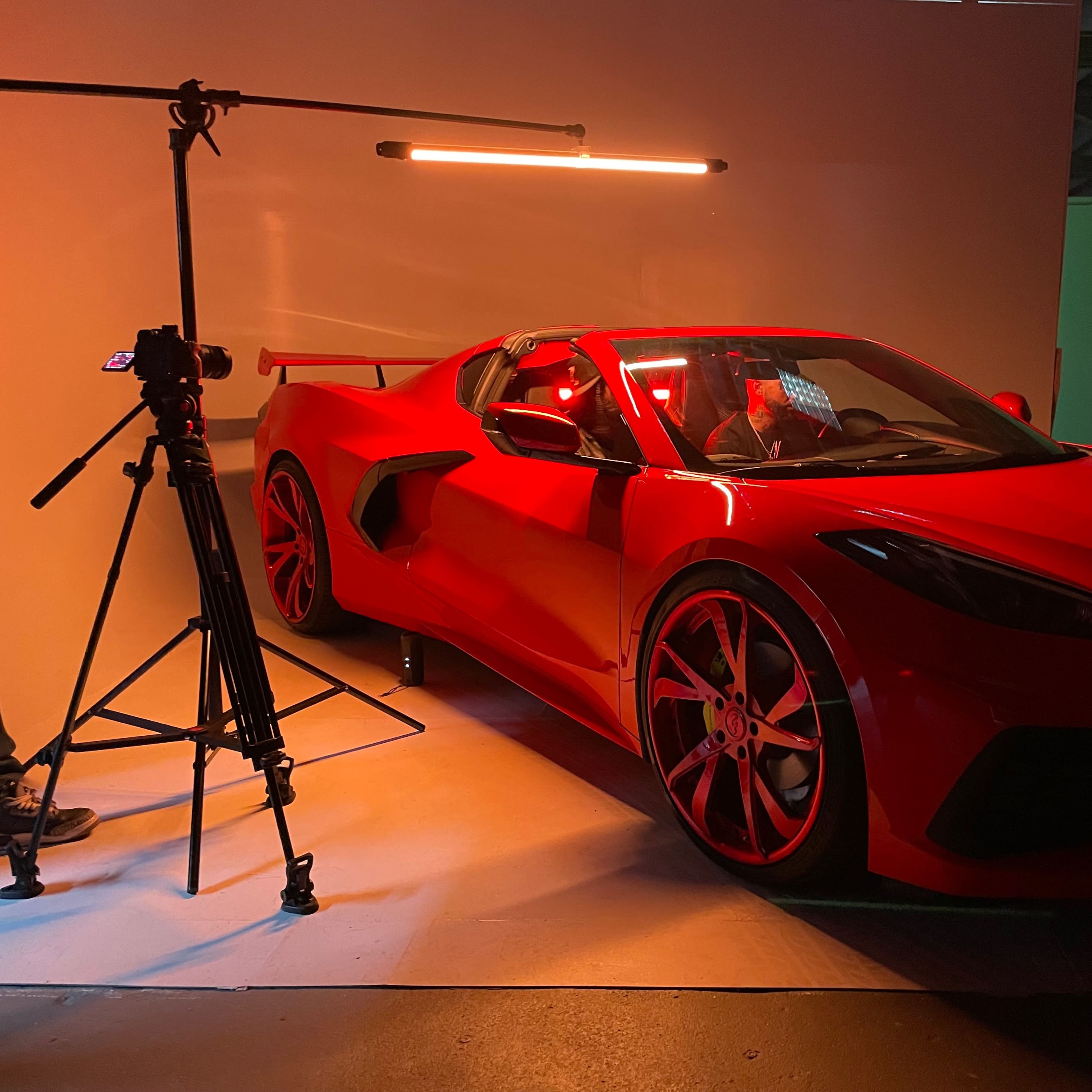 A red sports car is parked in a studio setting next to a photography tripod and light.