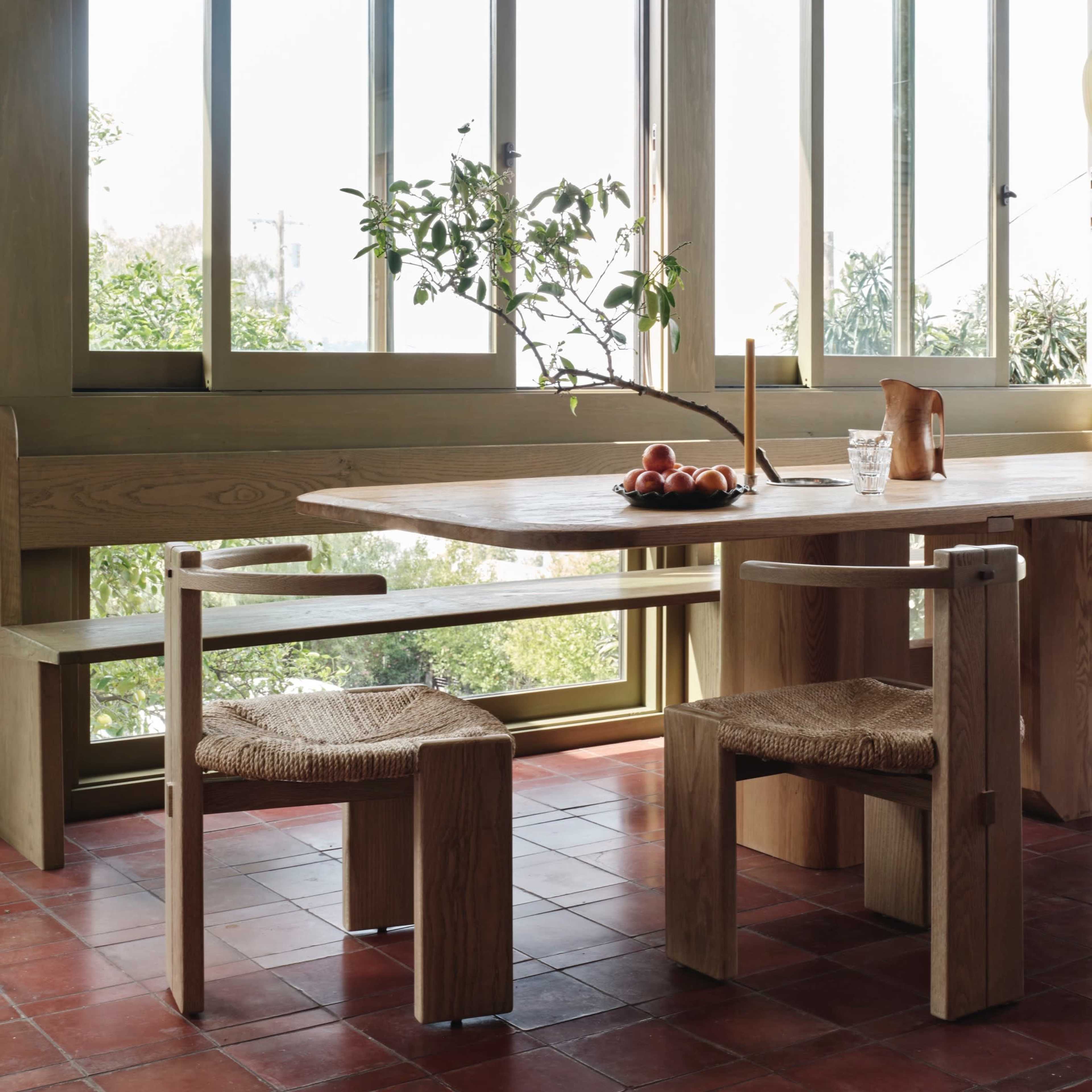 A wooden dining table with two chairs is set near large windows that overlook a garden.