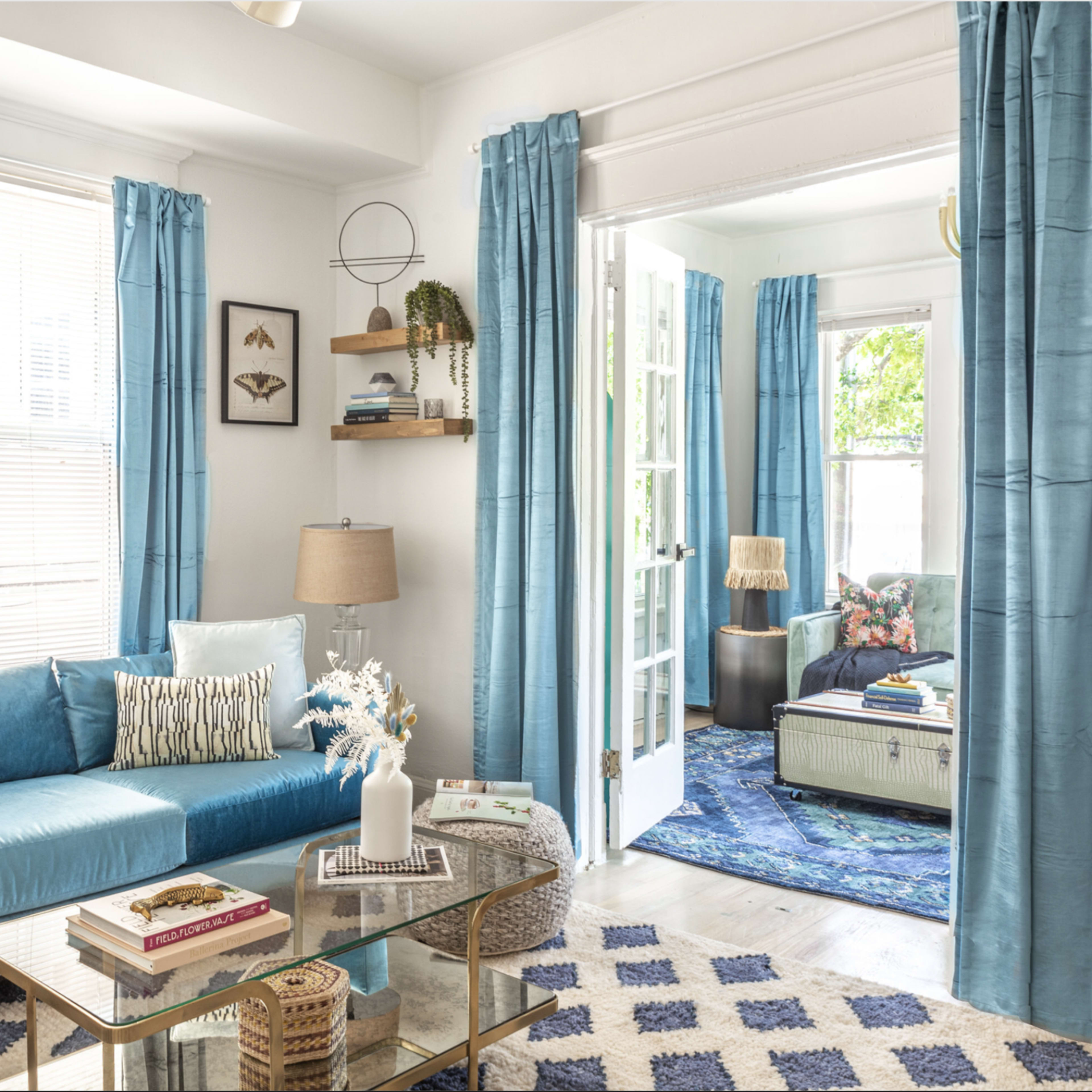 A living room features a blue sofa, light blue curtains, a glass coffee table, and a doorway leading to another room with floral decor.