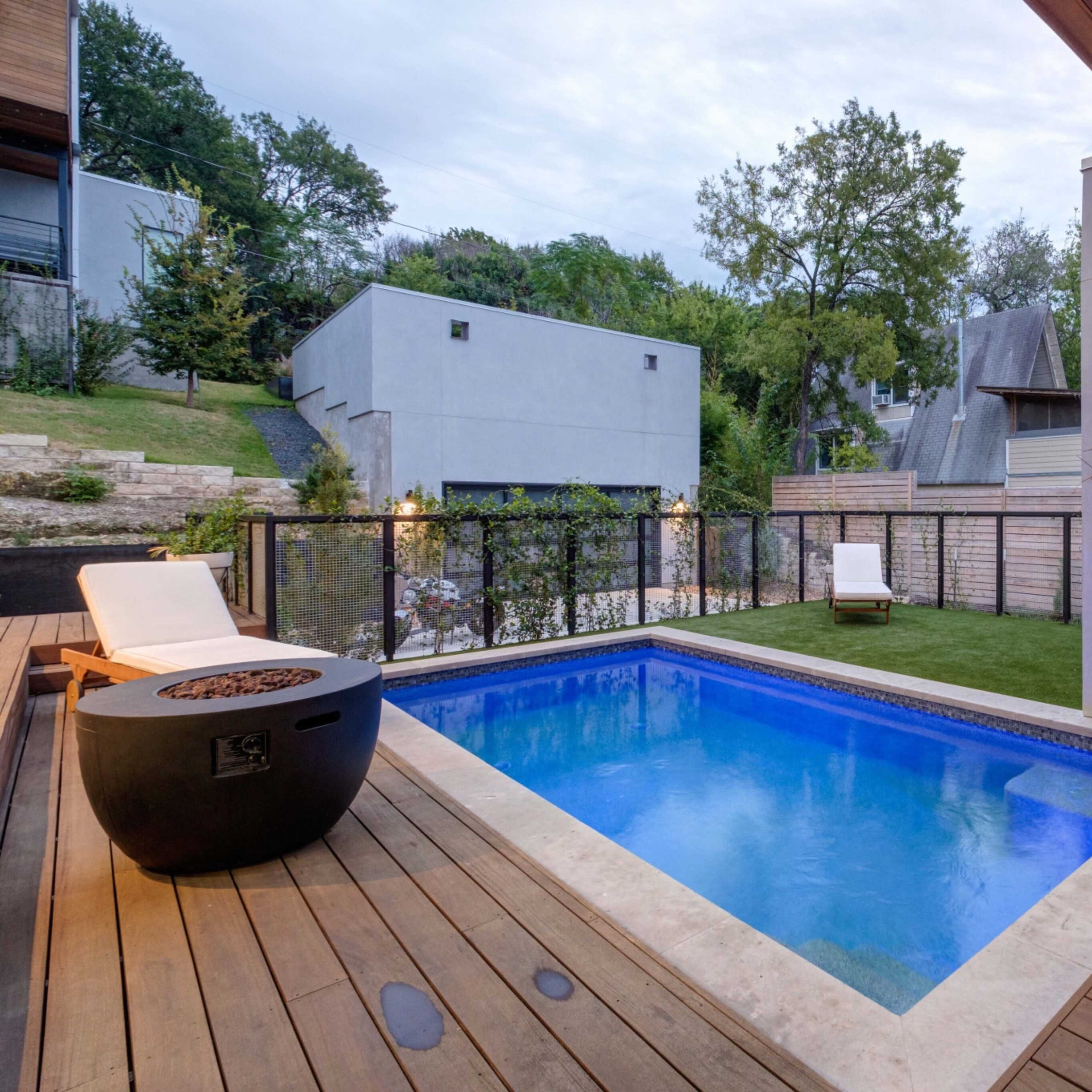 A modern, rectangular swimming pool surrounded by wooden decking and green grass, with a minimalist seating area and a backdrop of trees and buildings.