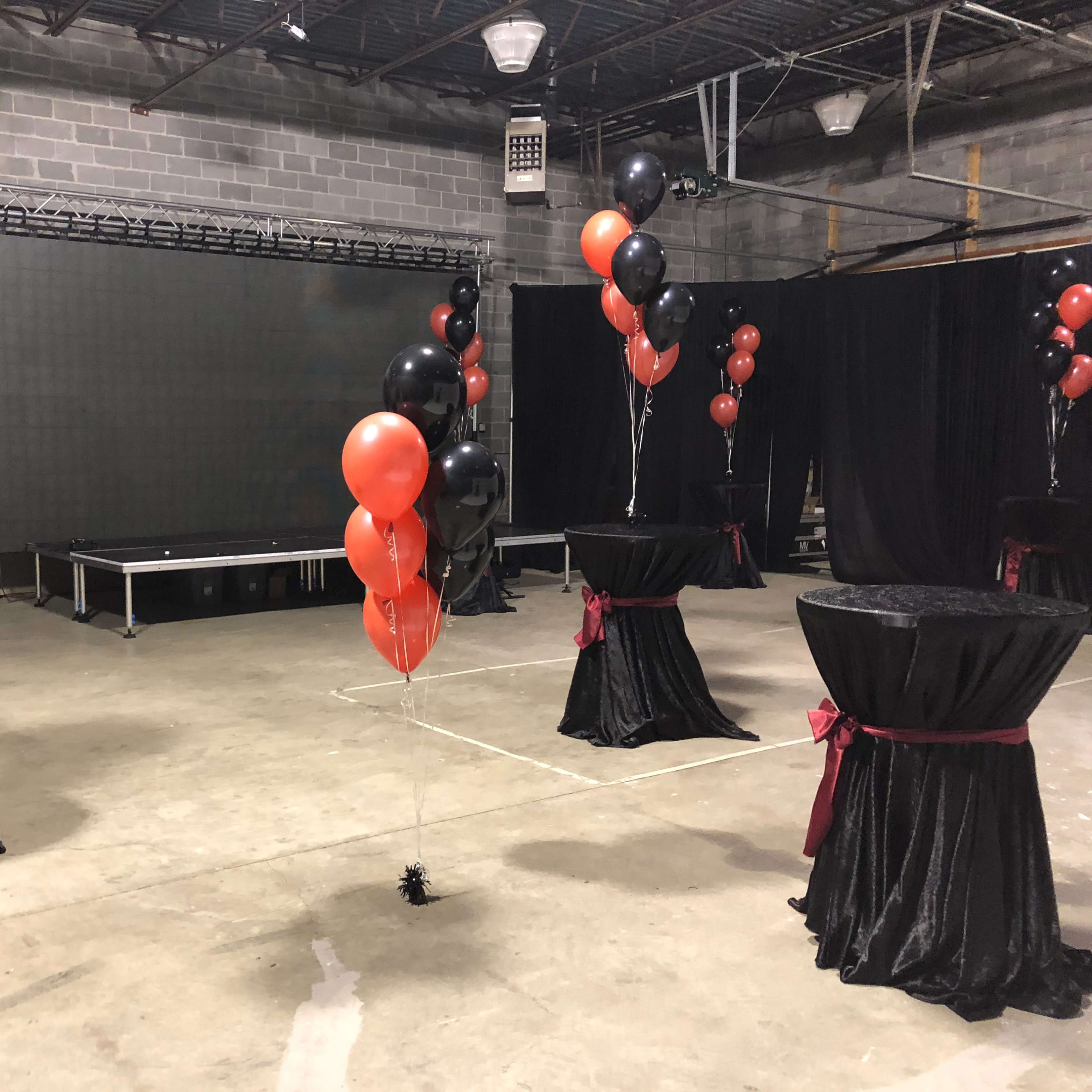 A decorated venue with black and red balloons on tables, a stage in the background, and black curtains.
