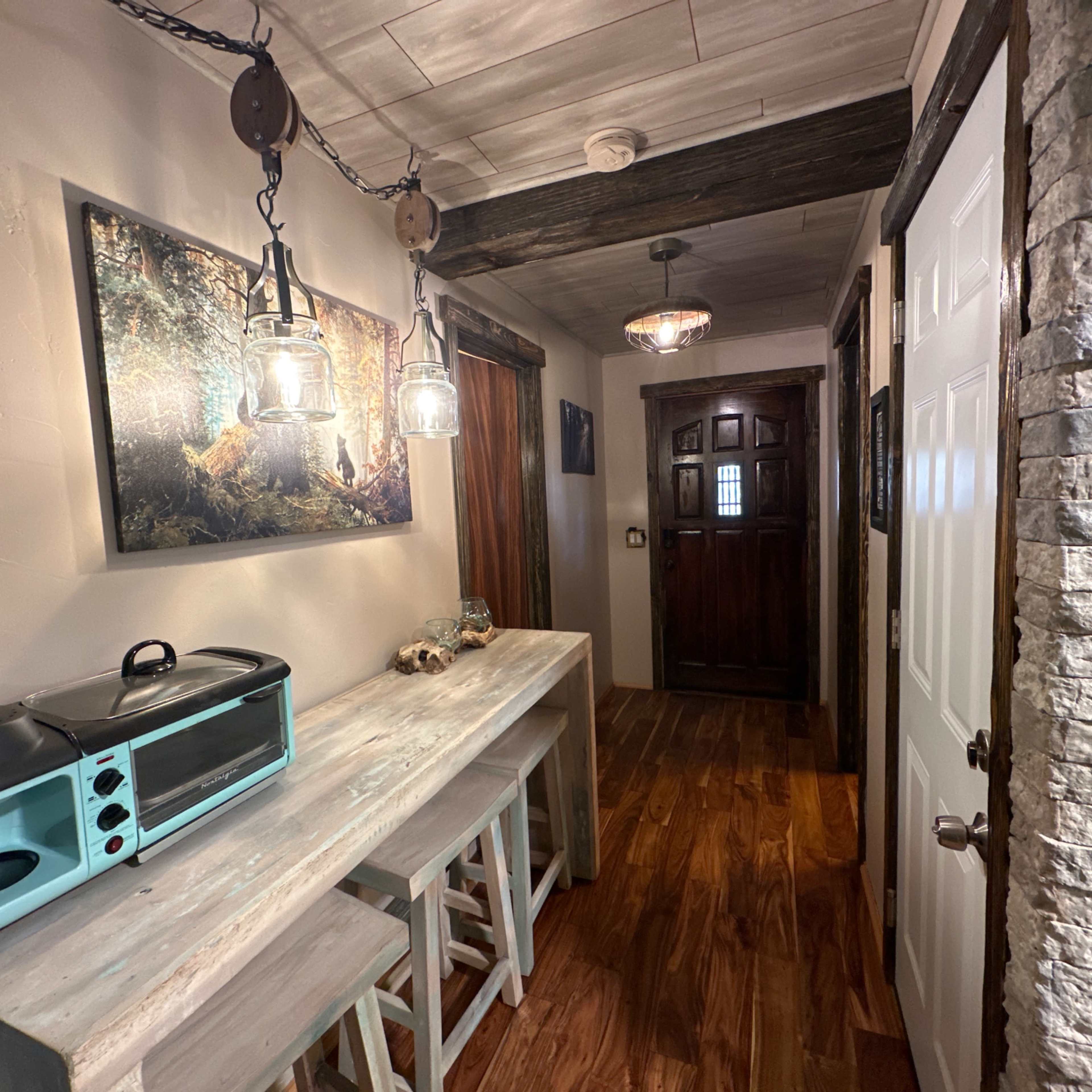 A narrow hallway with wooden flooring, a light-colored table with stools, and various light fixtures, leading to a wooden door at the end.