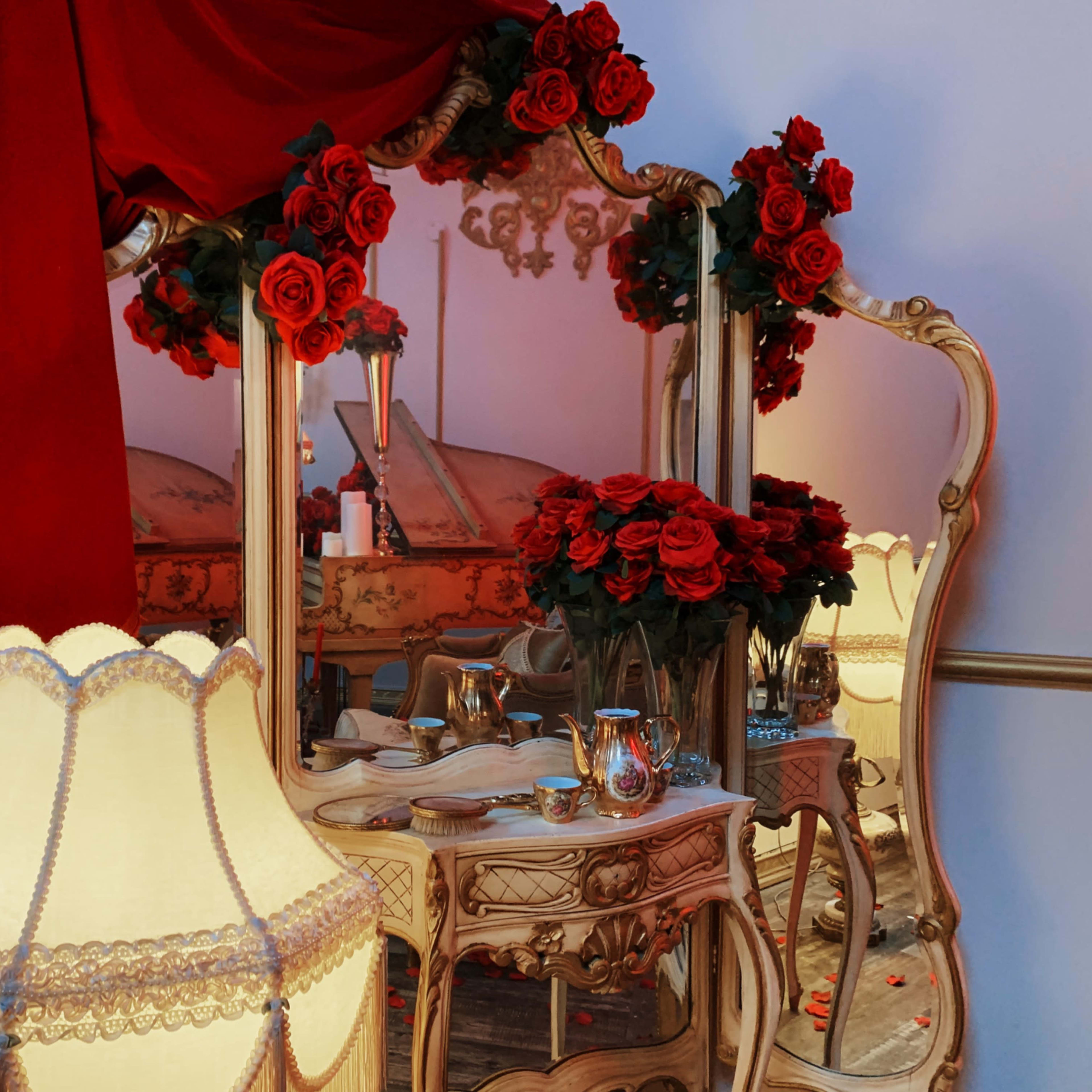 An ornate mirror surrounded by red roses and a vintage lamp, set against a backdrop of flowing red fabric.
