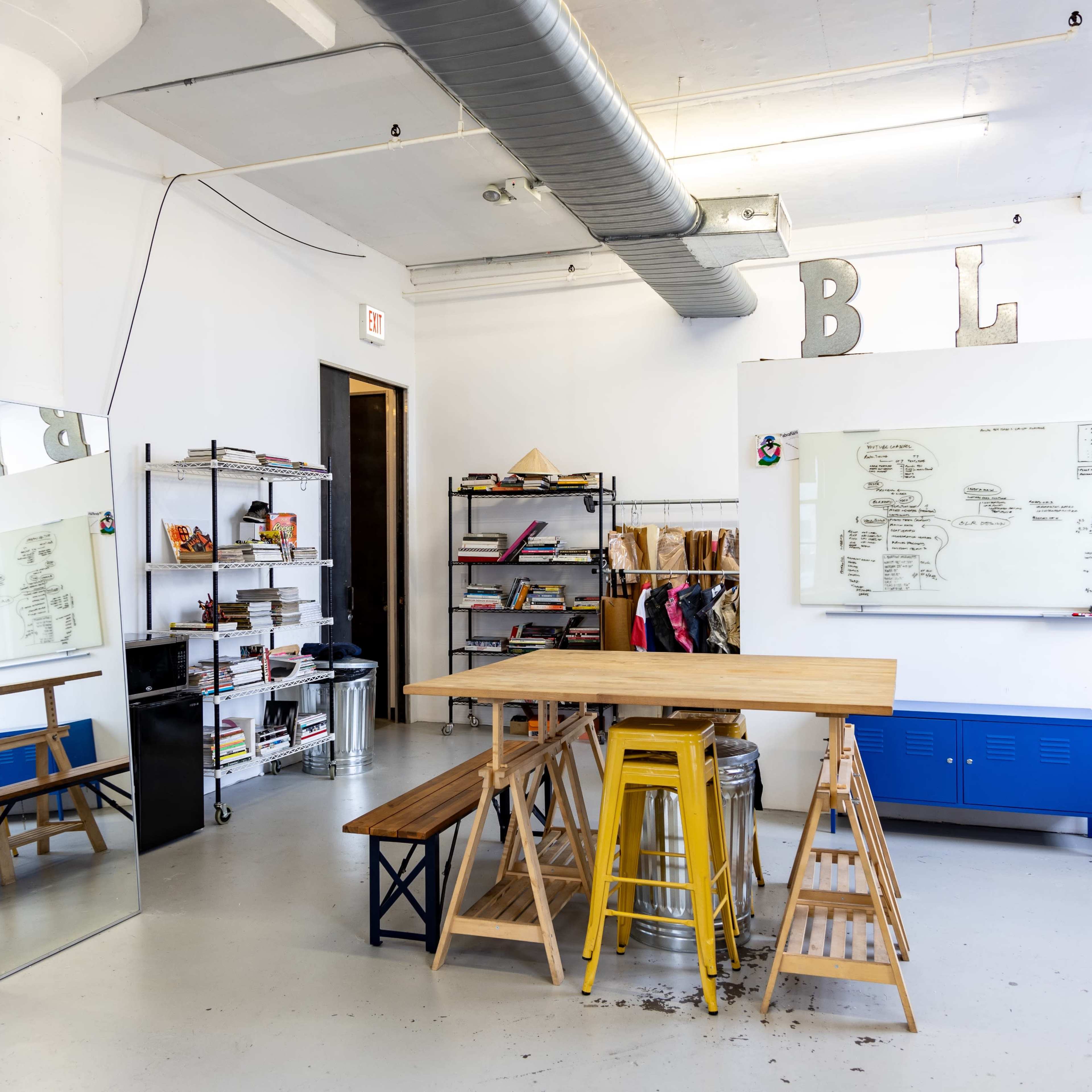 A bright, spacious room features a large table made of wooden planks, metal stools, a mirror, shelves filled with books and materials, and a whiteboard on the wall.
