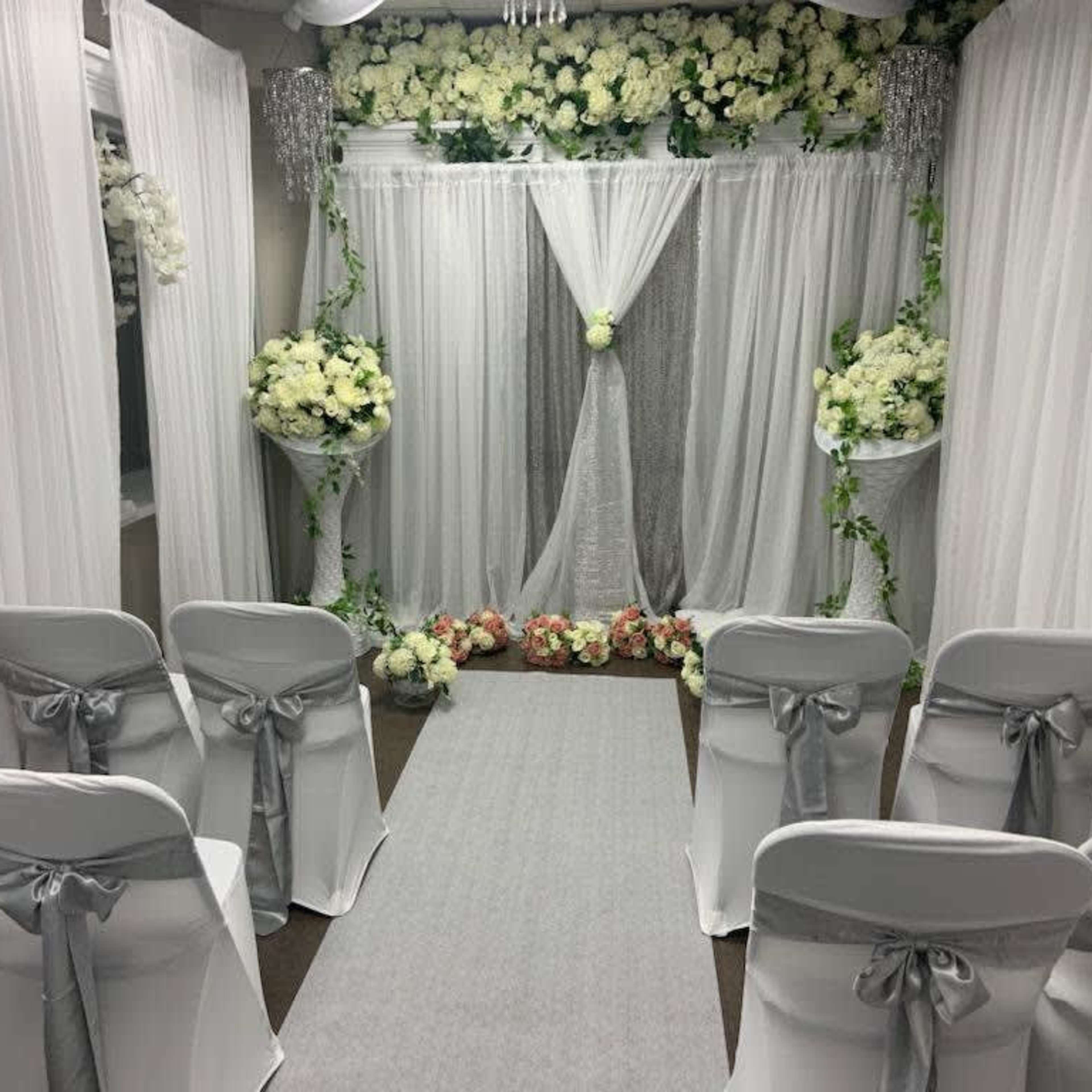 A wedding ceremony setup with white drapes, floral arrangements, and chairs arranged along a gray aisle.