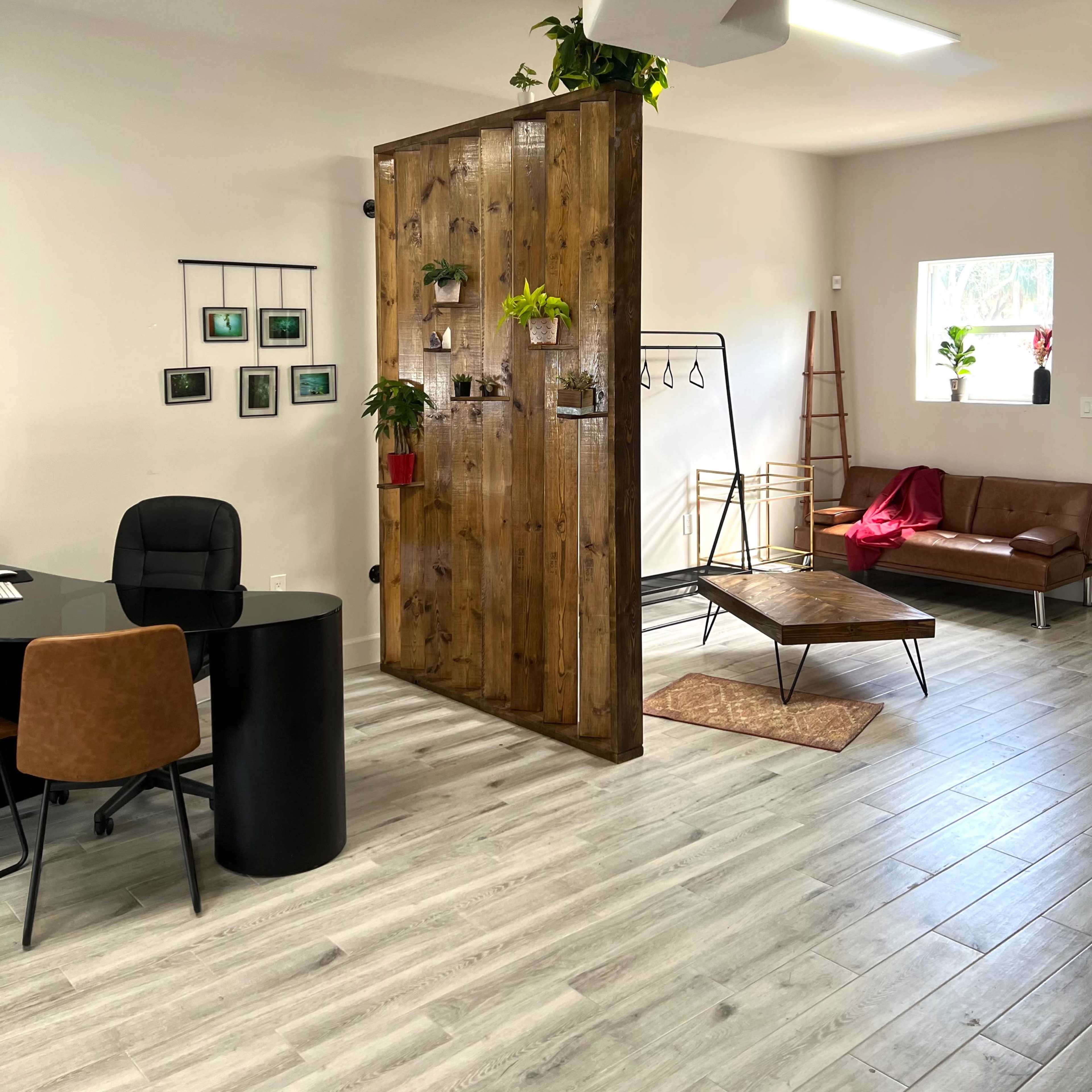 A modern office space with a wooden partition, a black desk and chair, a brown leather couch, and plants integrated into the decor.