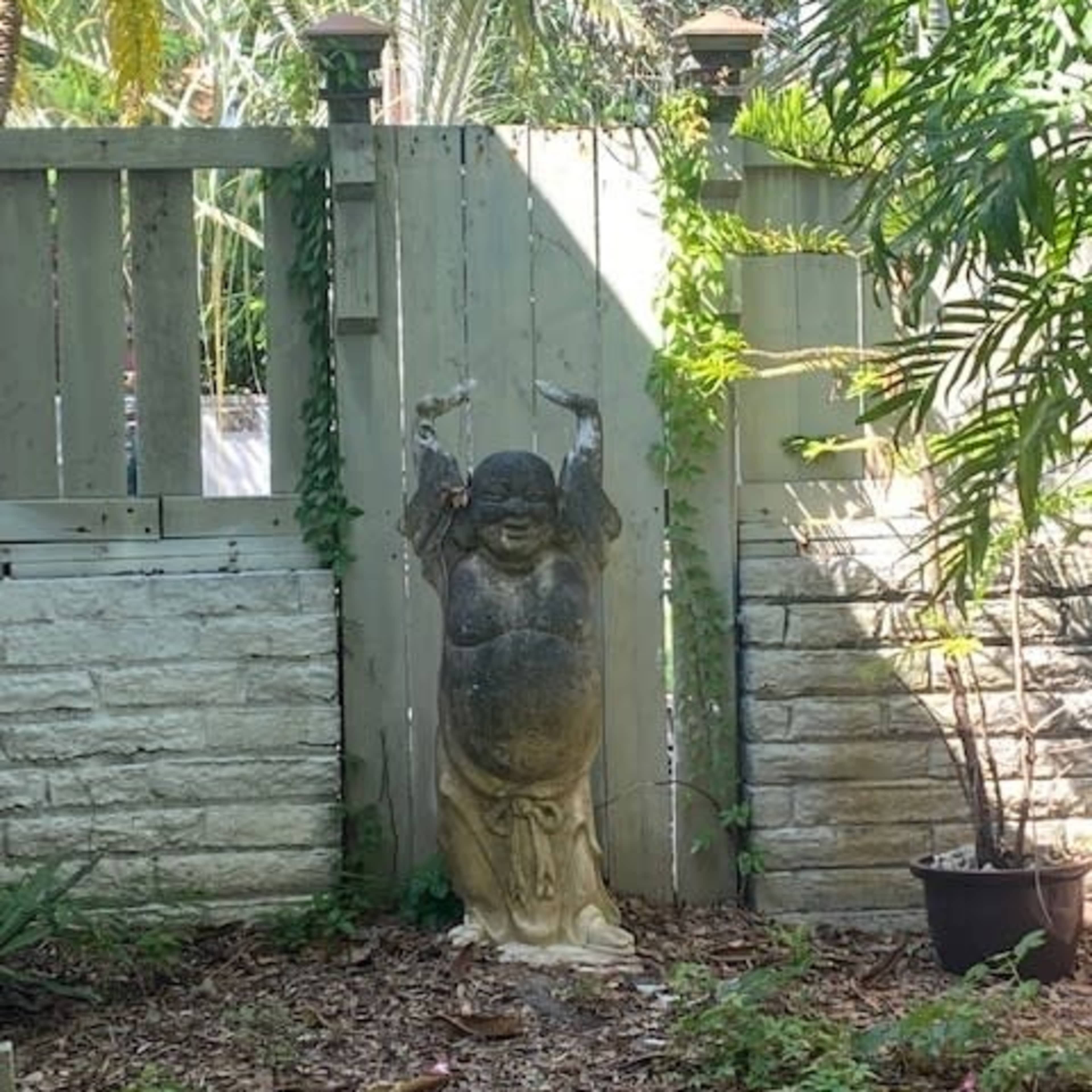 A stone statue of a smiling figure with raised arms stands beside a wooden fence in a garden with tropical plants.