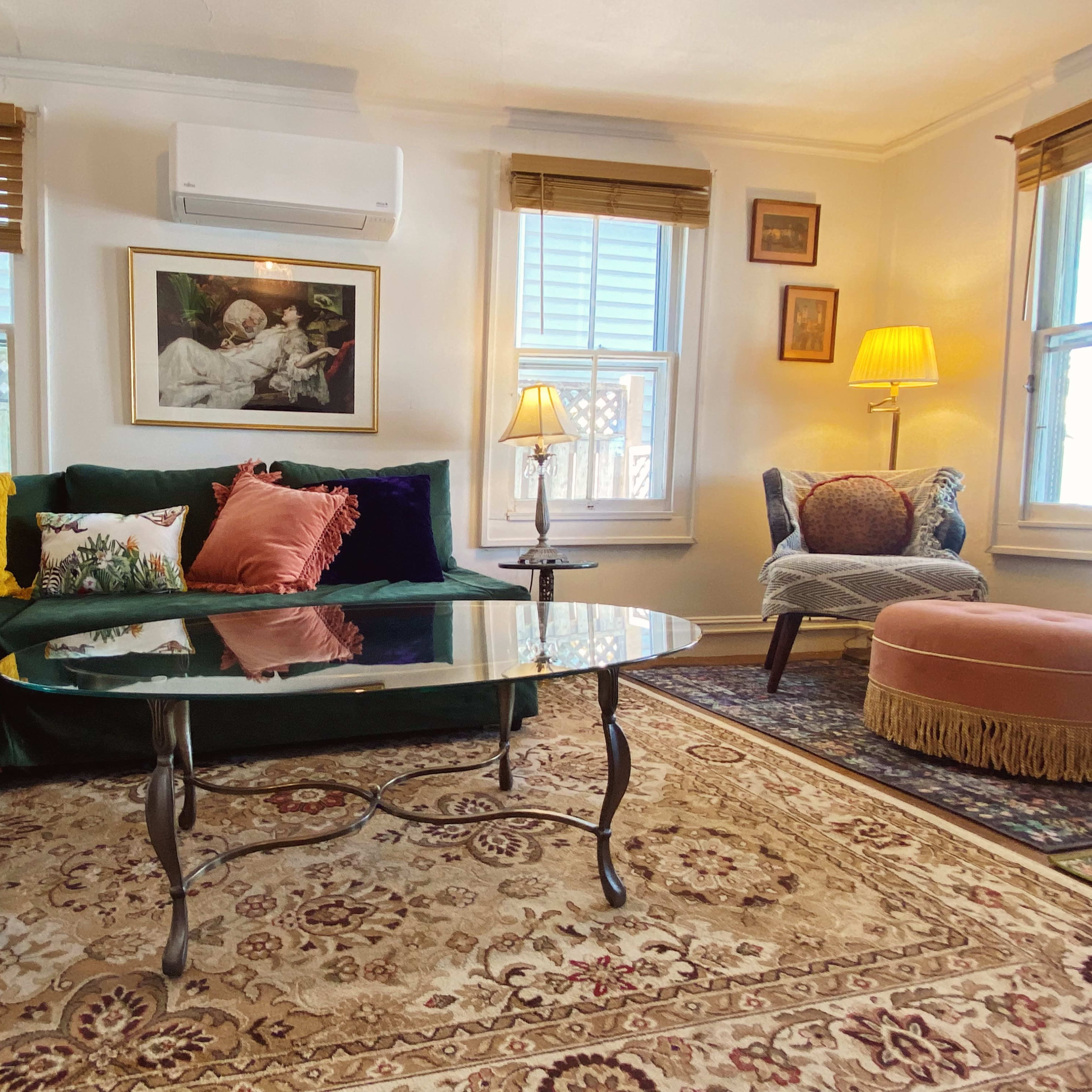 A cozy living room featuring a green sectional sofa adorned with colorful pillows, a glass coffee table, and a patterned rug, with ample natural light coming through two windows.