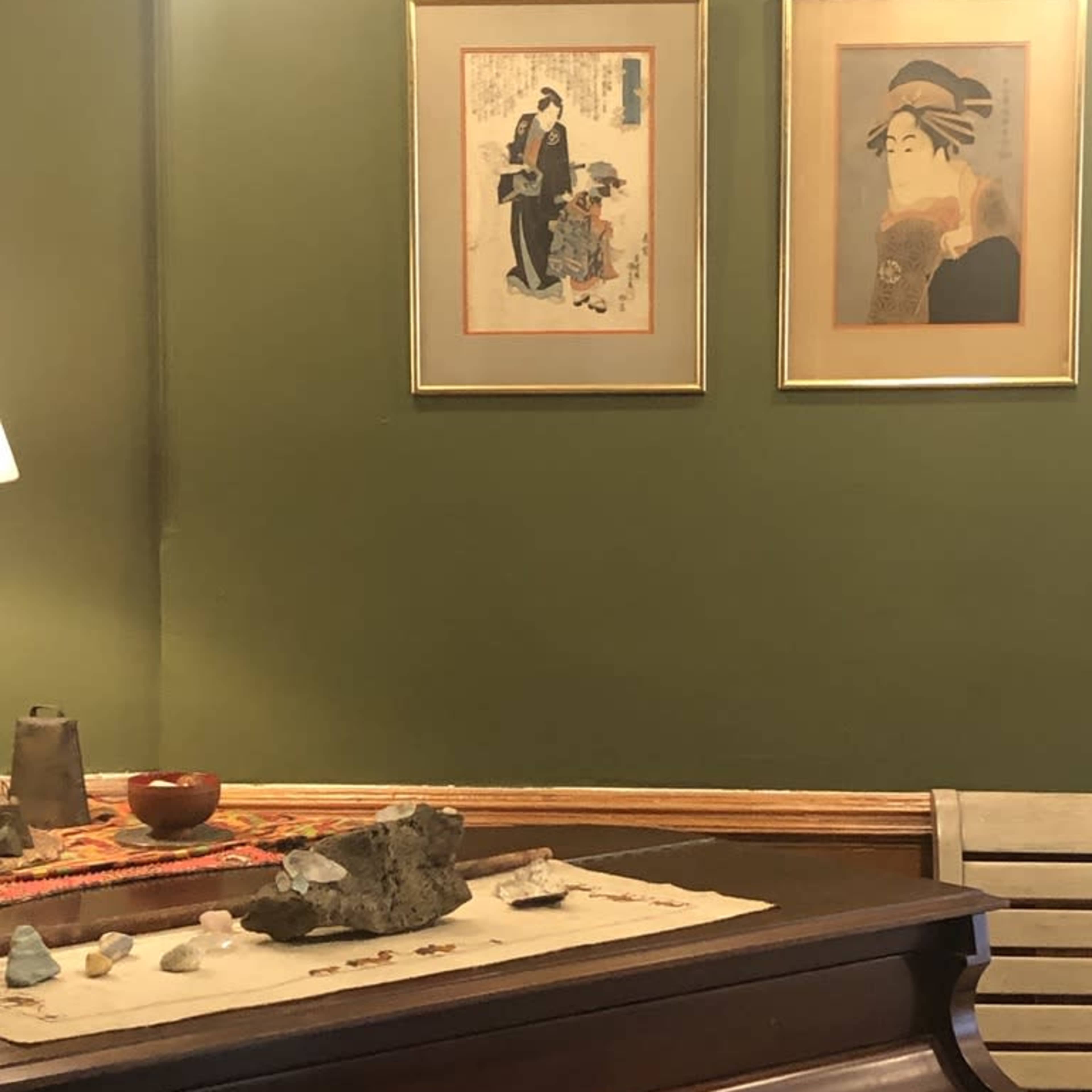 A wooden piano is placed against a green wall, adorned with two framed artworks and a decorative table displaying various small objects.