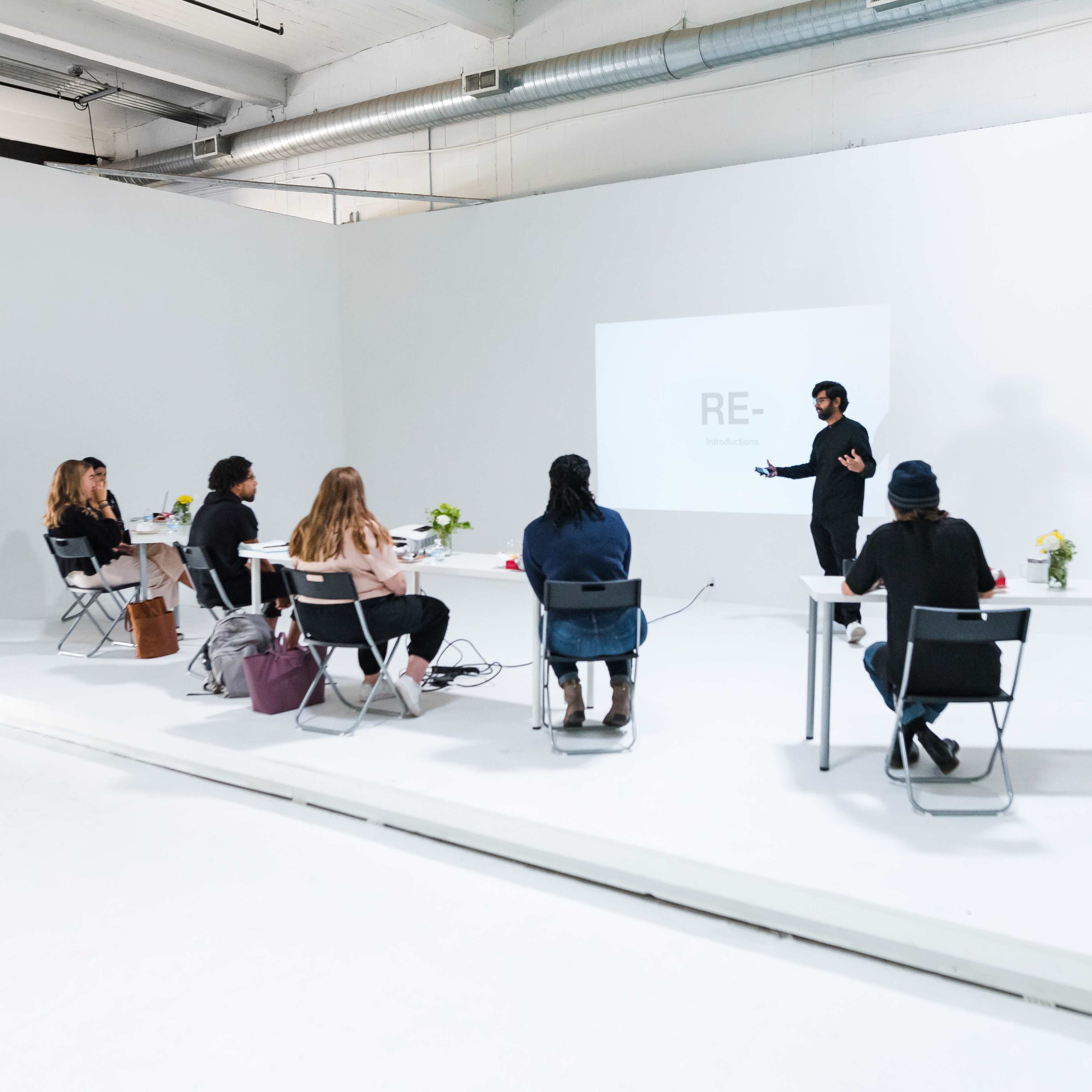 A group of people is seated at tables in a minimalist room, where a presenter is speaking in front of a projection screen.