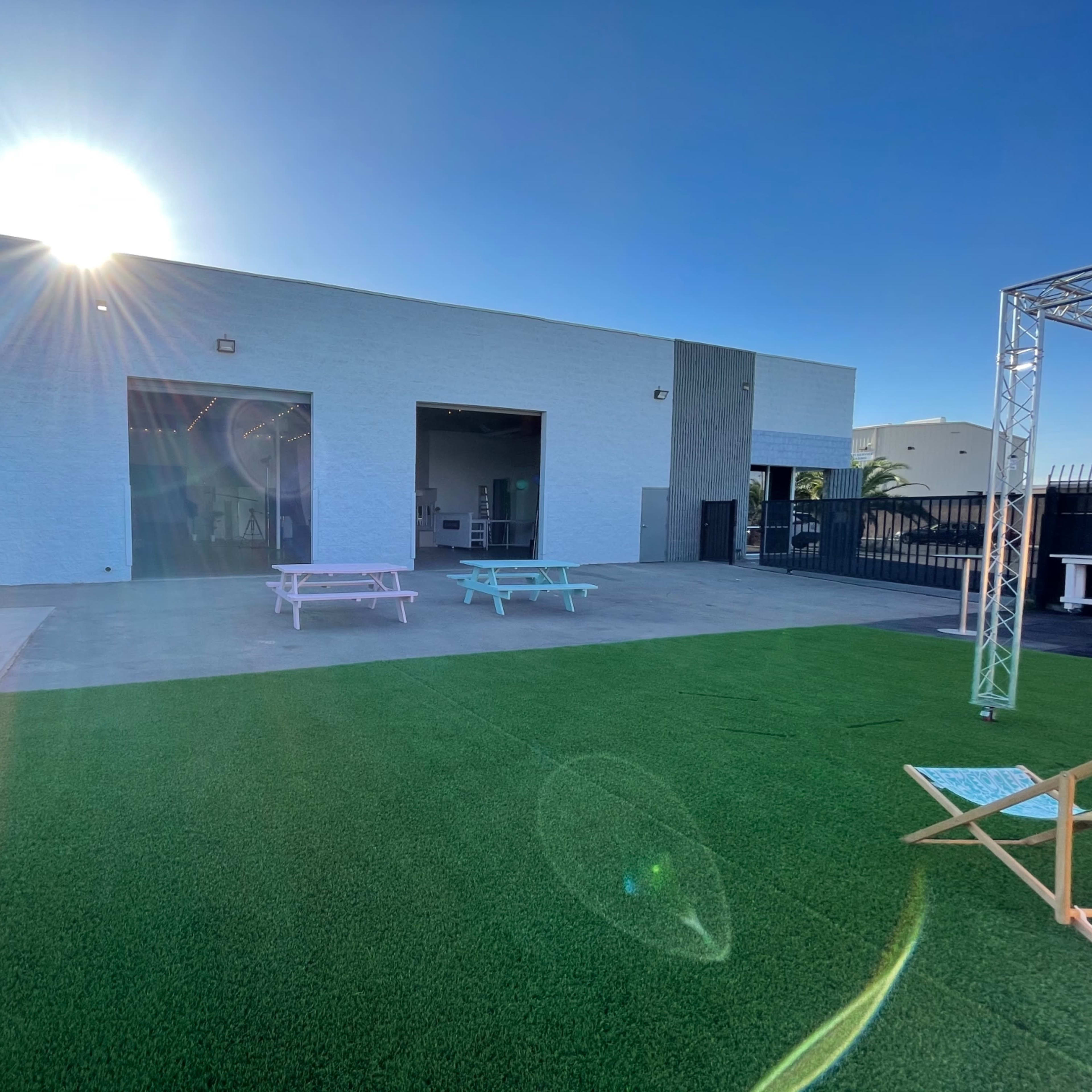 A light-colored building with large doors is set against a clear blue sky, with green artificial grass and patio furniture in the foreground.
