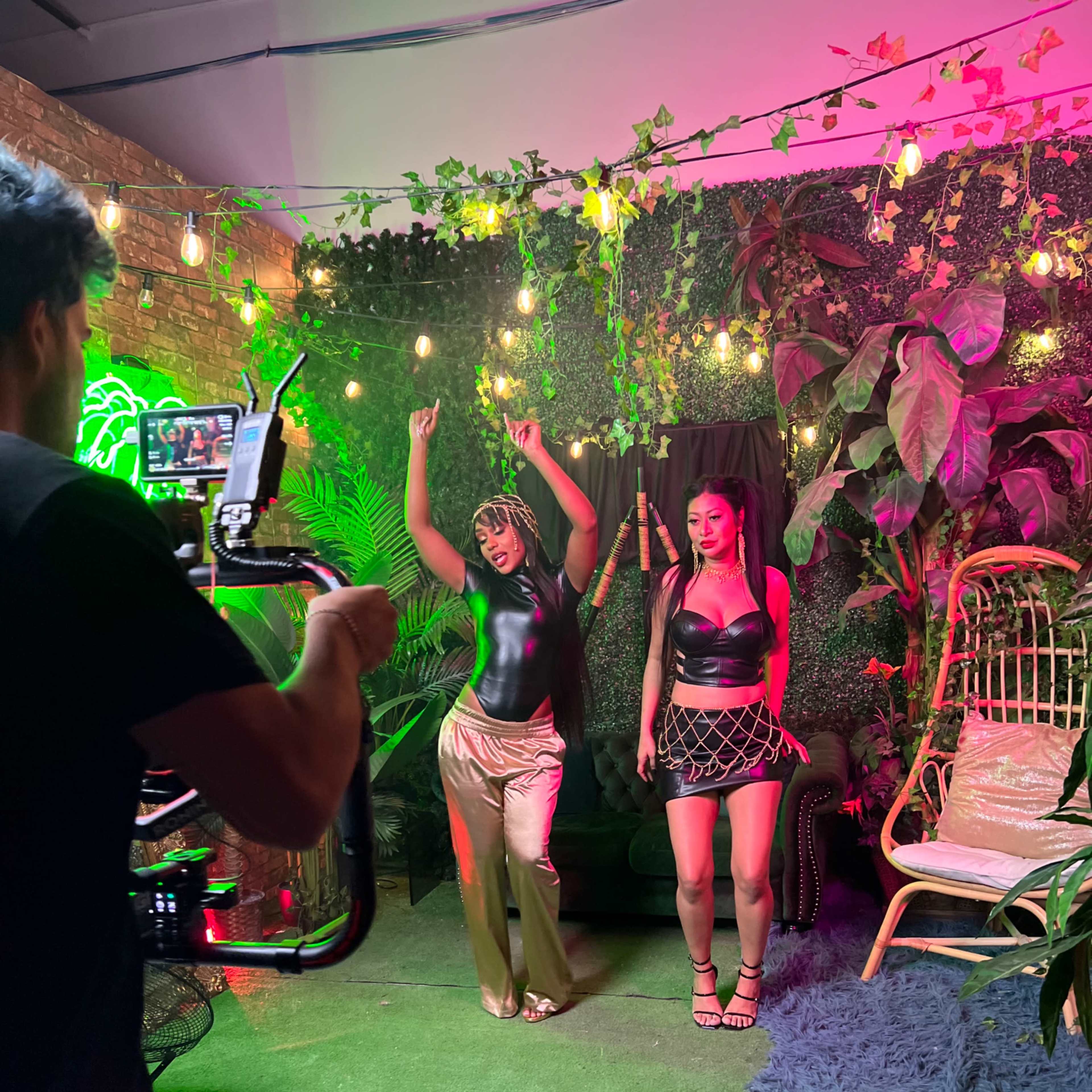 Two dancers perform in a studio decorated with plants and colorful lights while being filmed by a person holding a camera.