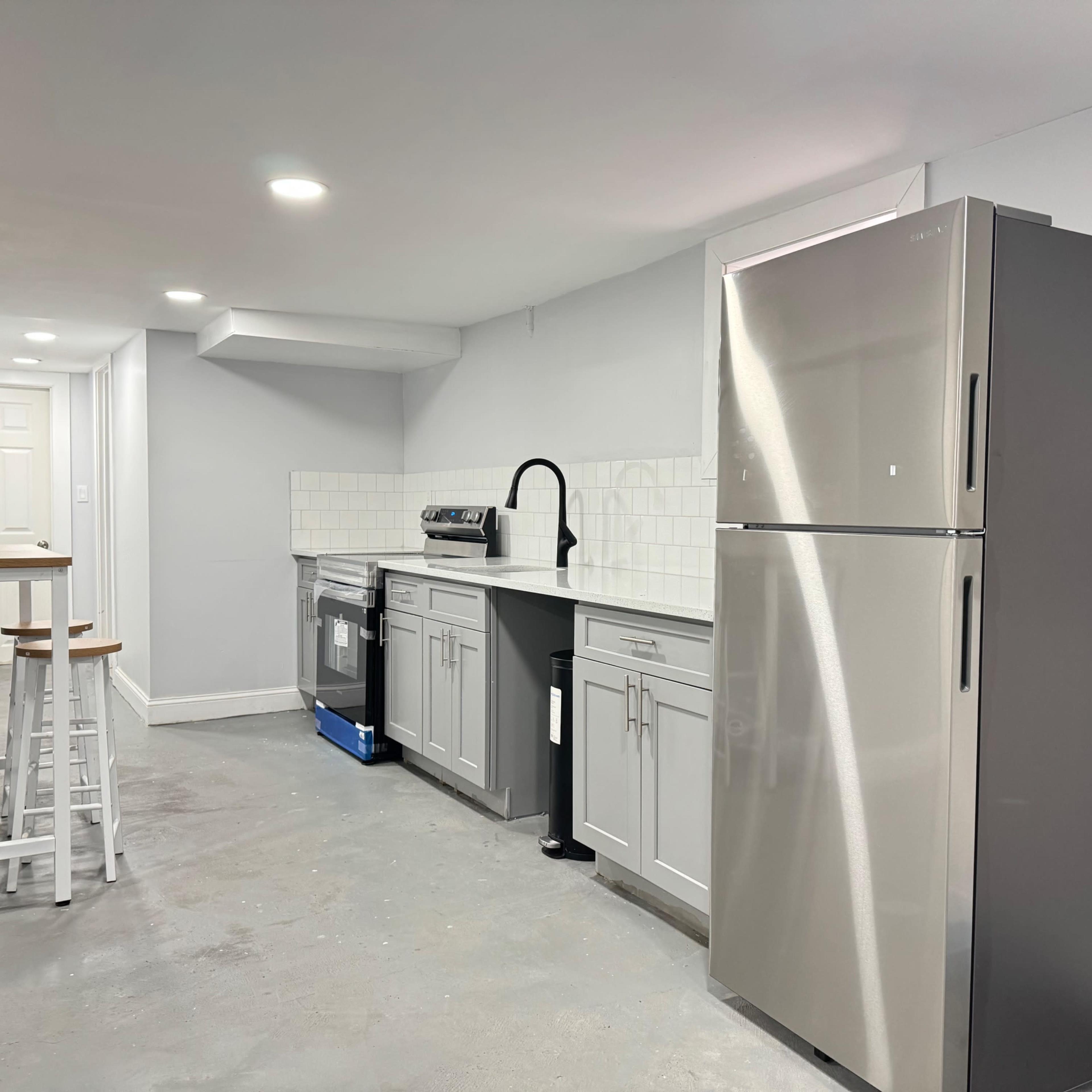 A modern kitchen area with gray cabinets, a stainless steel refrigerator, and a small table with stools against the wall.