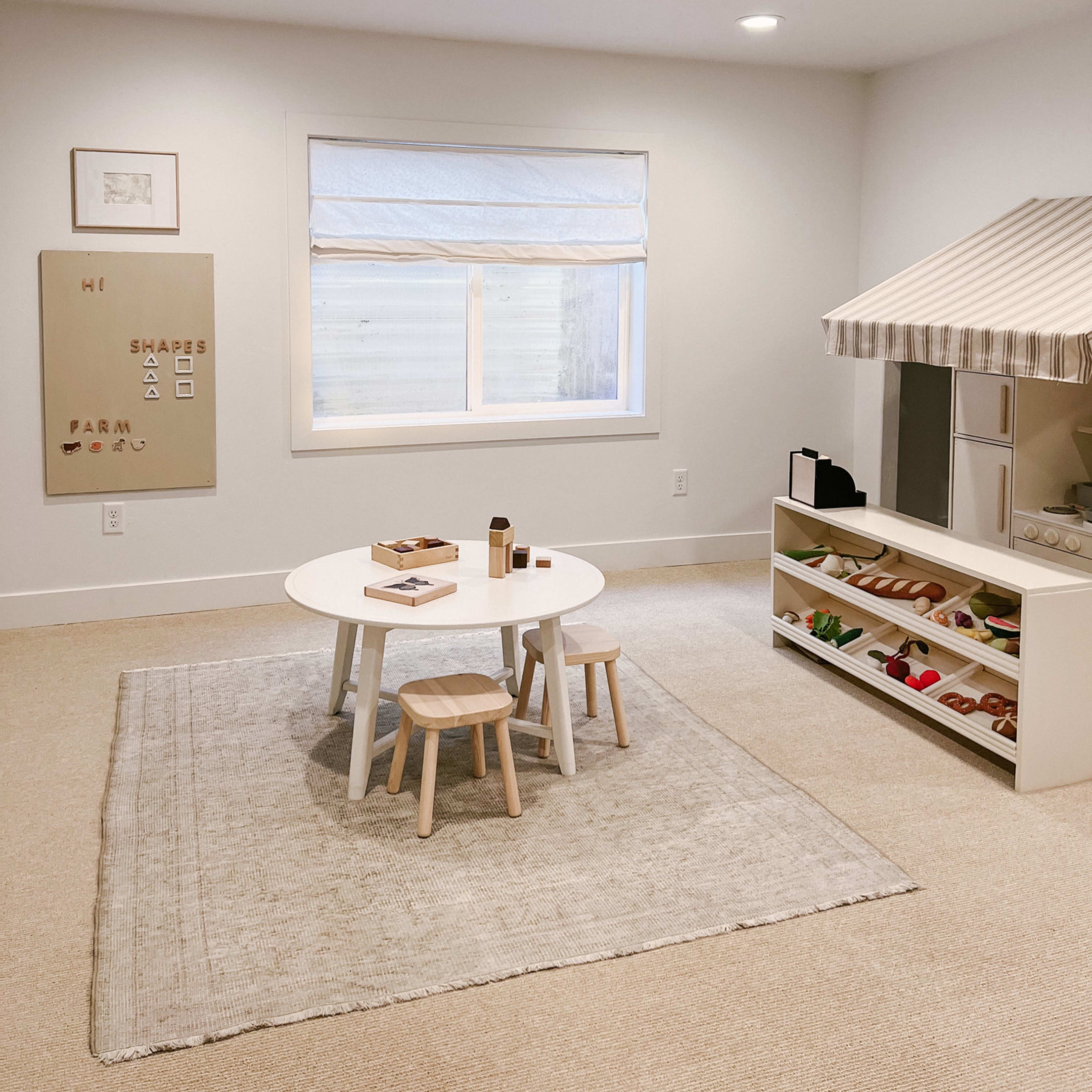 A minimalist playroom with a circular table, two small stools, and a shelf filled with toys, featuring a plush rug and a window with a light-filtering treatment.