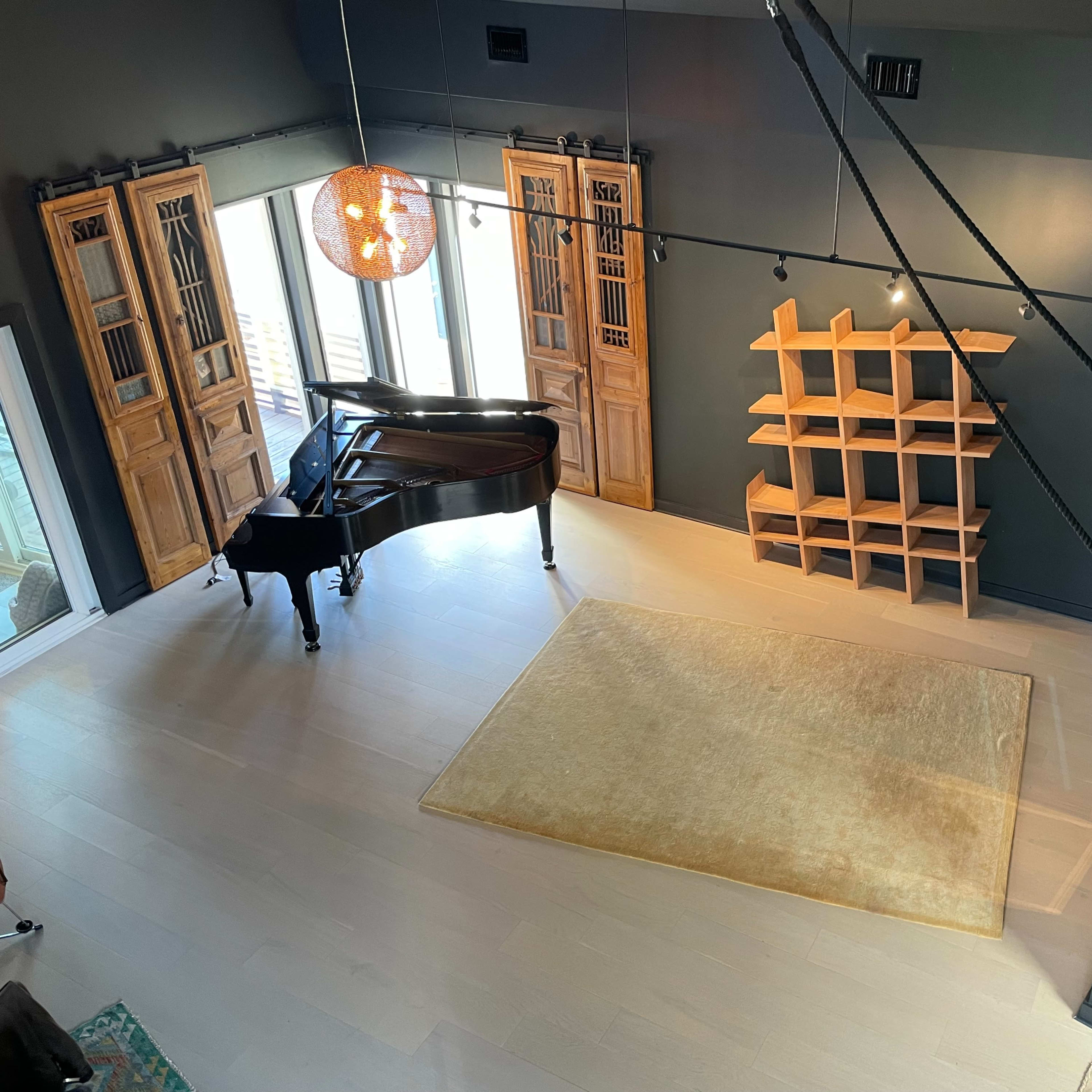 A spacious room with a grand piano, wooden storage units, and a large area rug on a light wooden floor, illuminated by a pendant light hanging from the ceiling.