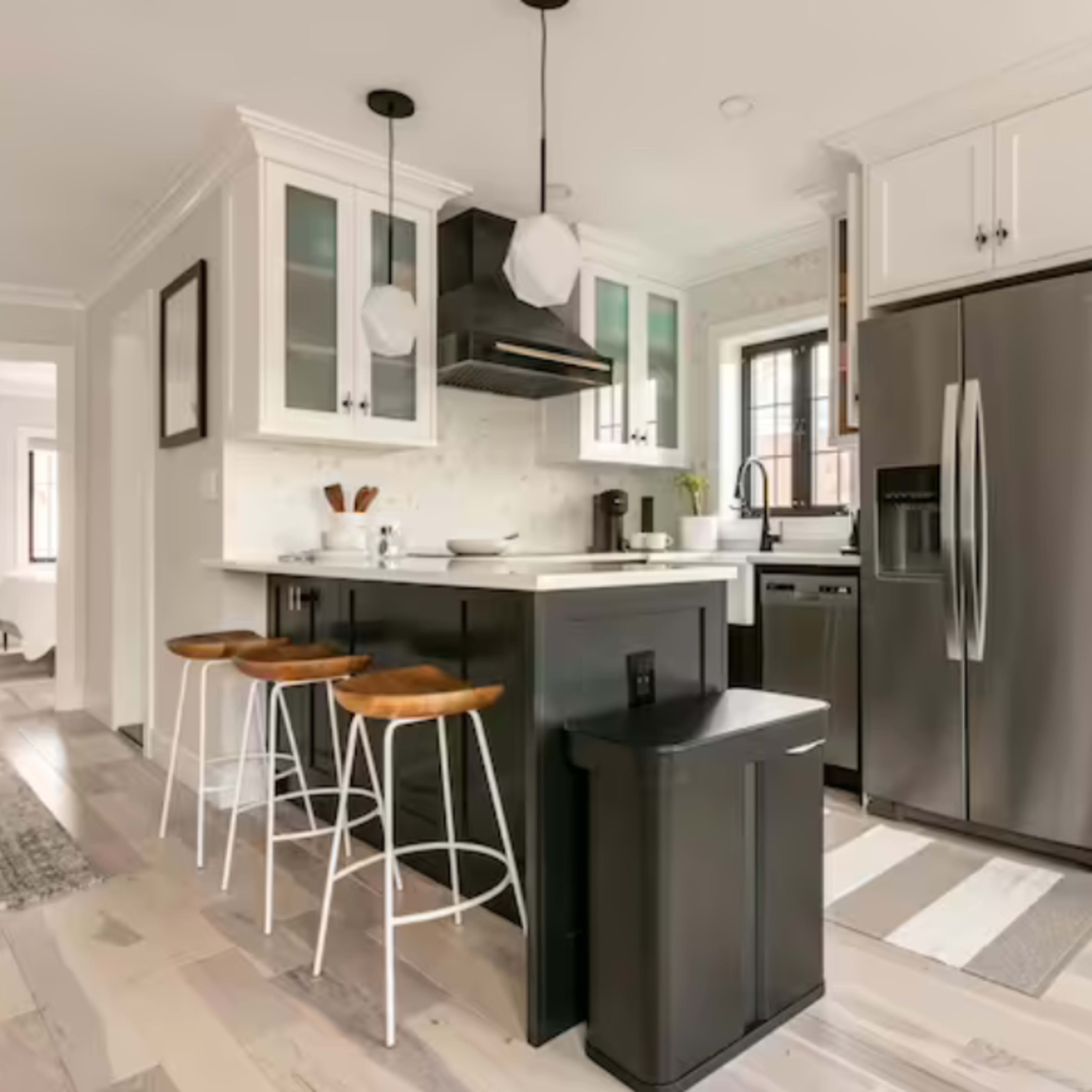 A modern kitchen features a central island with three stools, stainless steel appliances, and cabinetry with glass accents.