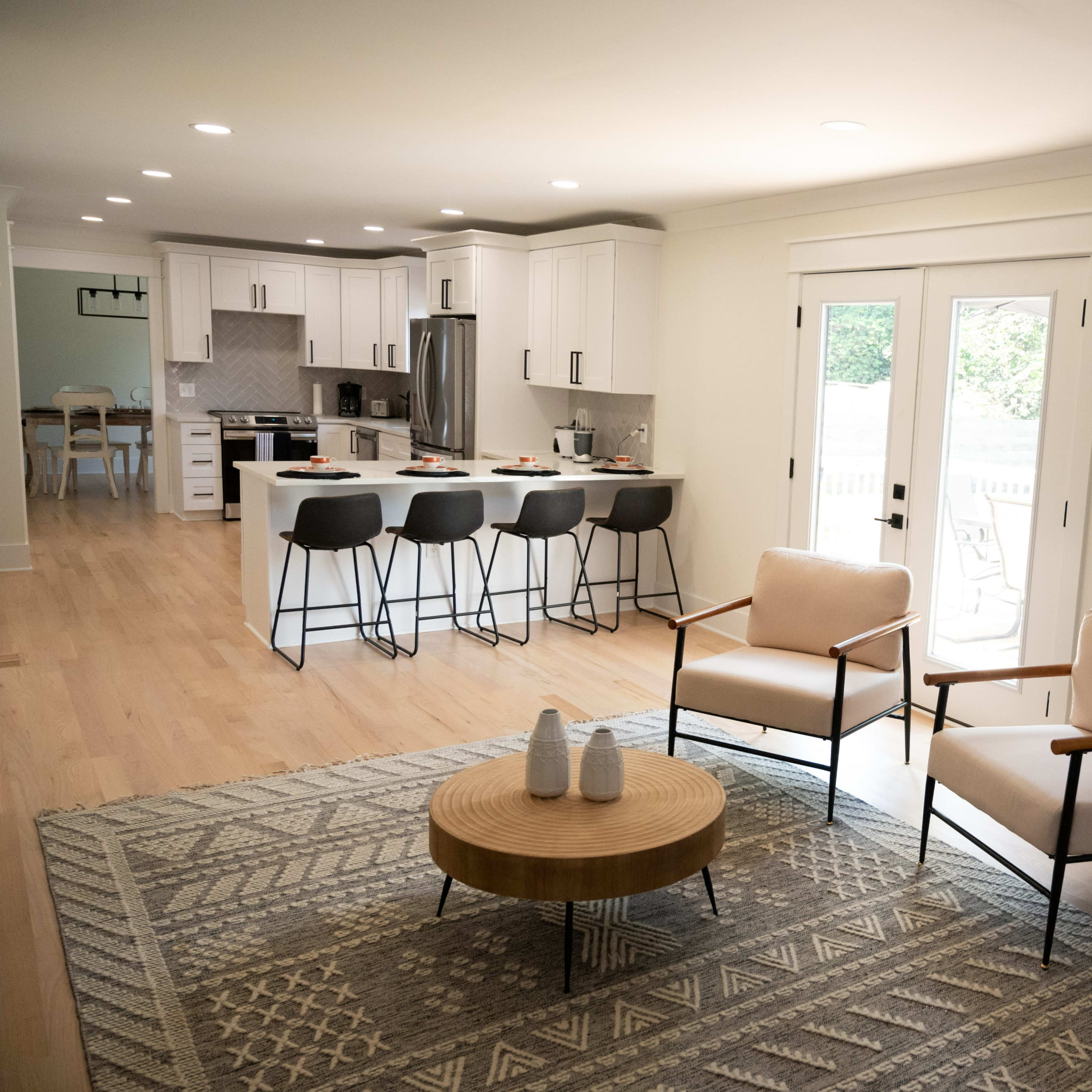 A modern living area features two armchairs and a circular coffee table with a geometric rug, adjacent to a kitchen with bar stools and white cabinetry.