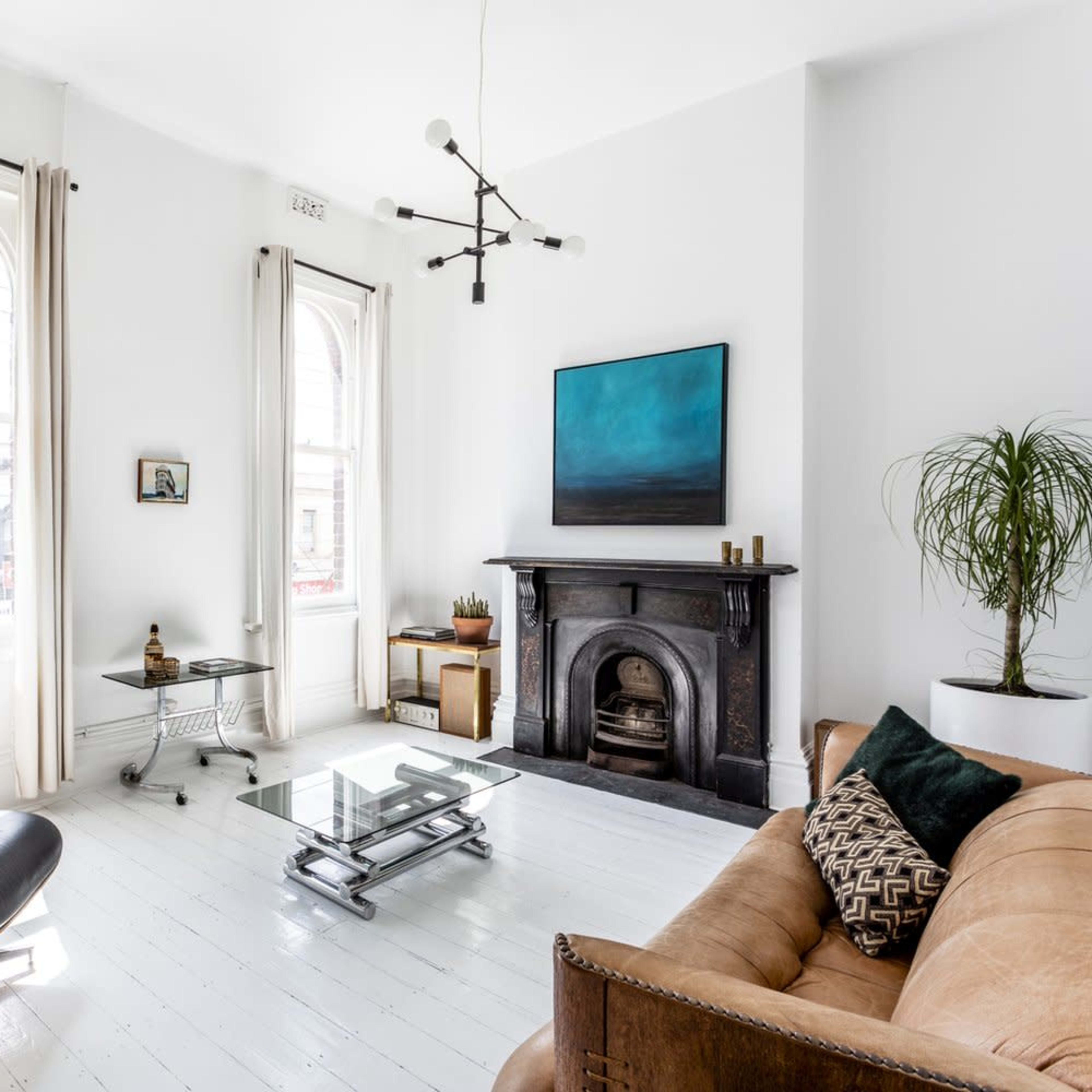 A modern living room features a leather sofa, an Eames chair, a glass coffee table, and a fireplace, with large windows allowing natural light to fill the space.