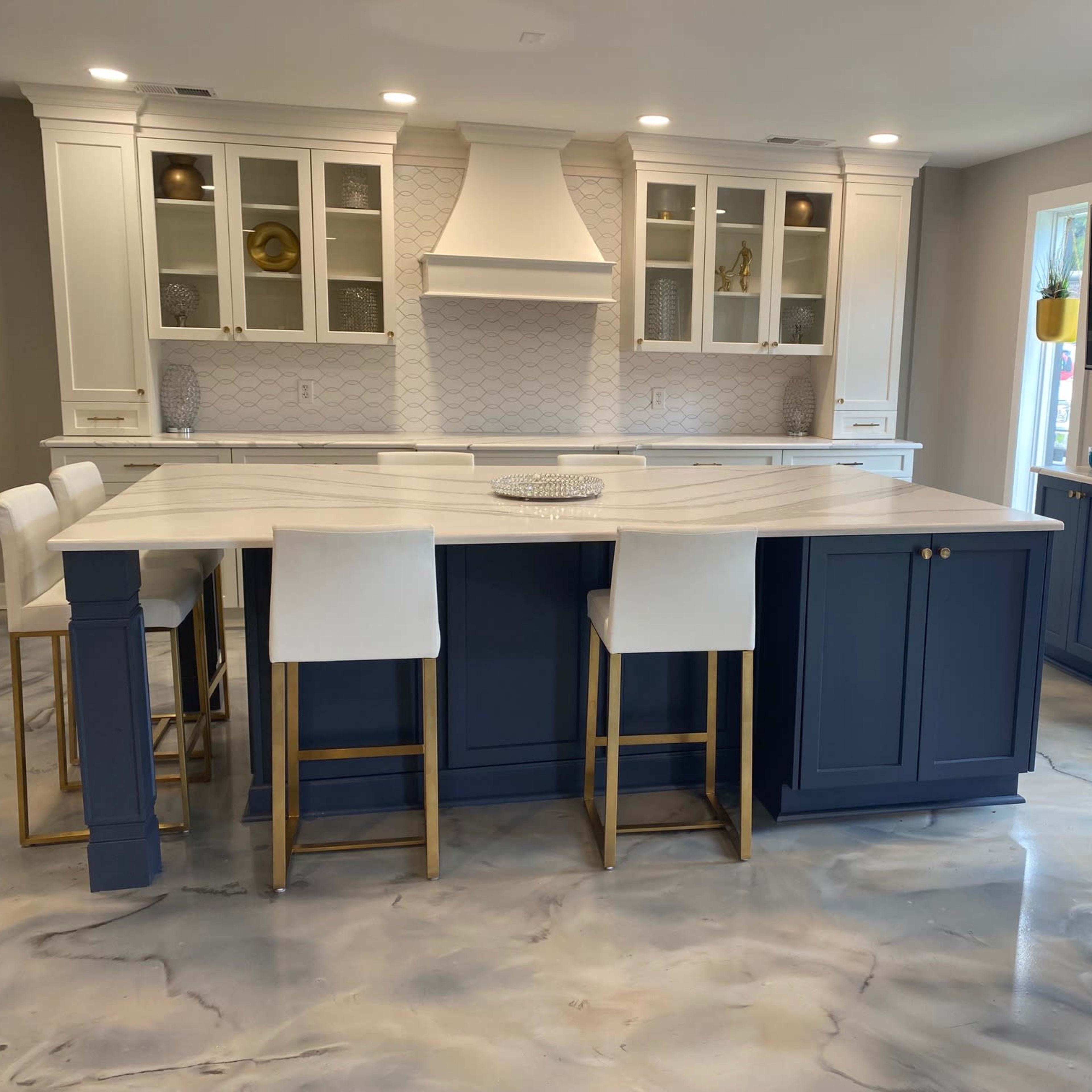 A modern kitchen featuring a large central island with white and gray marble countertops, navy blue cabinetry, and gold-accented bar stools.