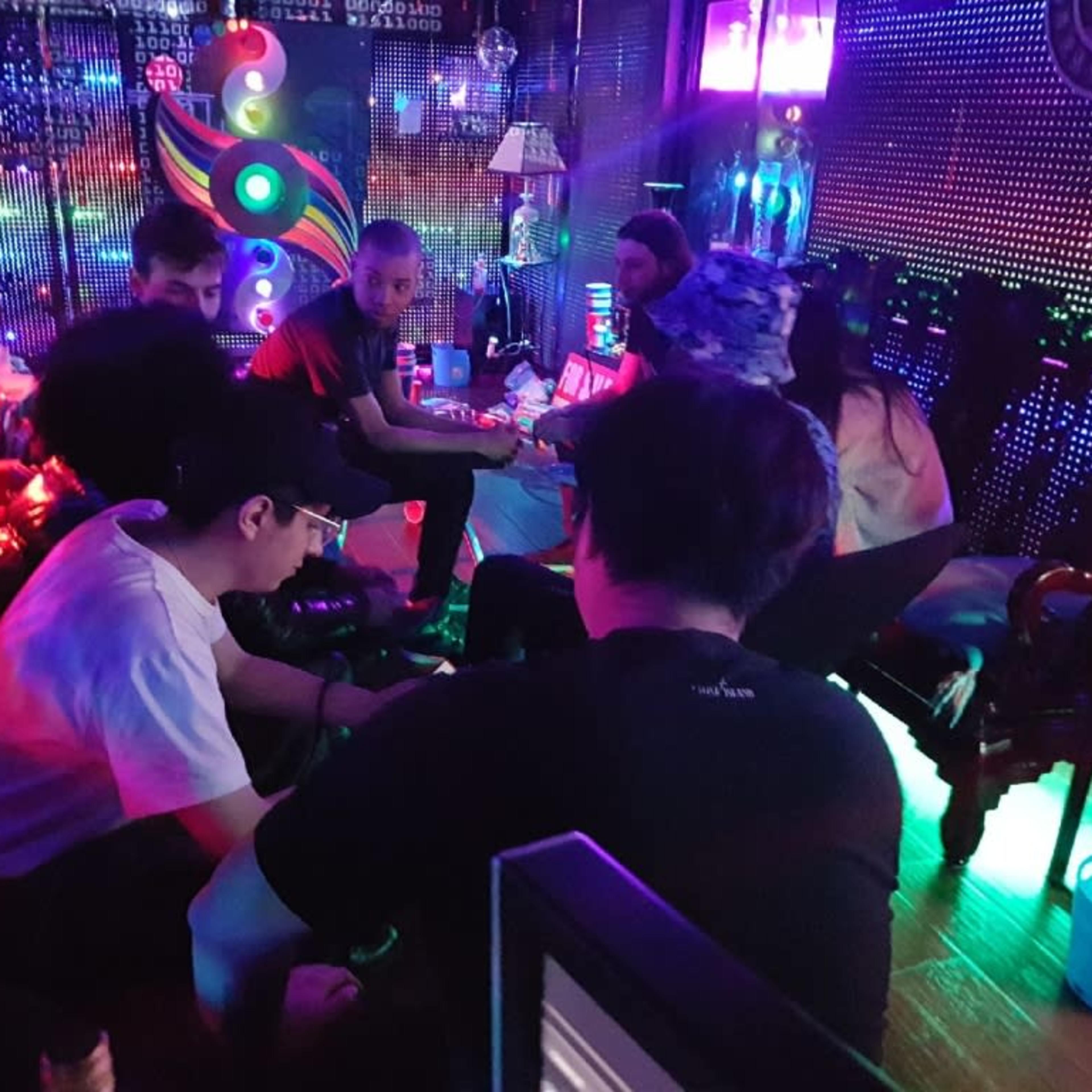 A group of people sits in a dimly lit room with colorful lights, engaged in conversation around a table.