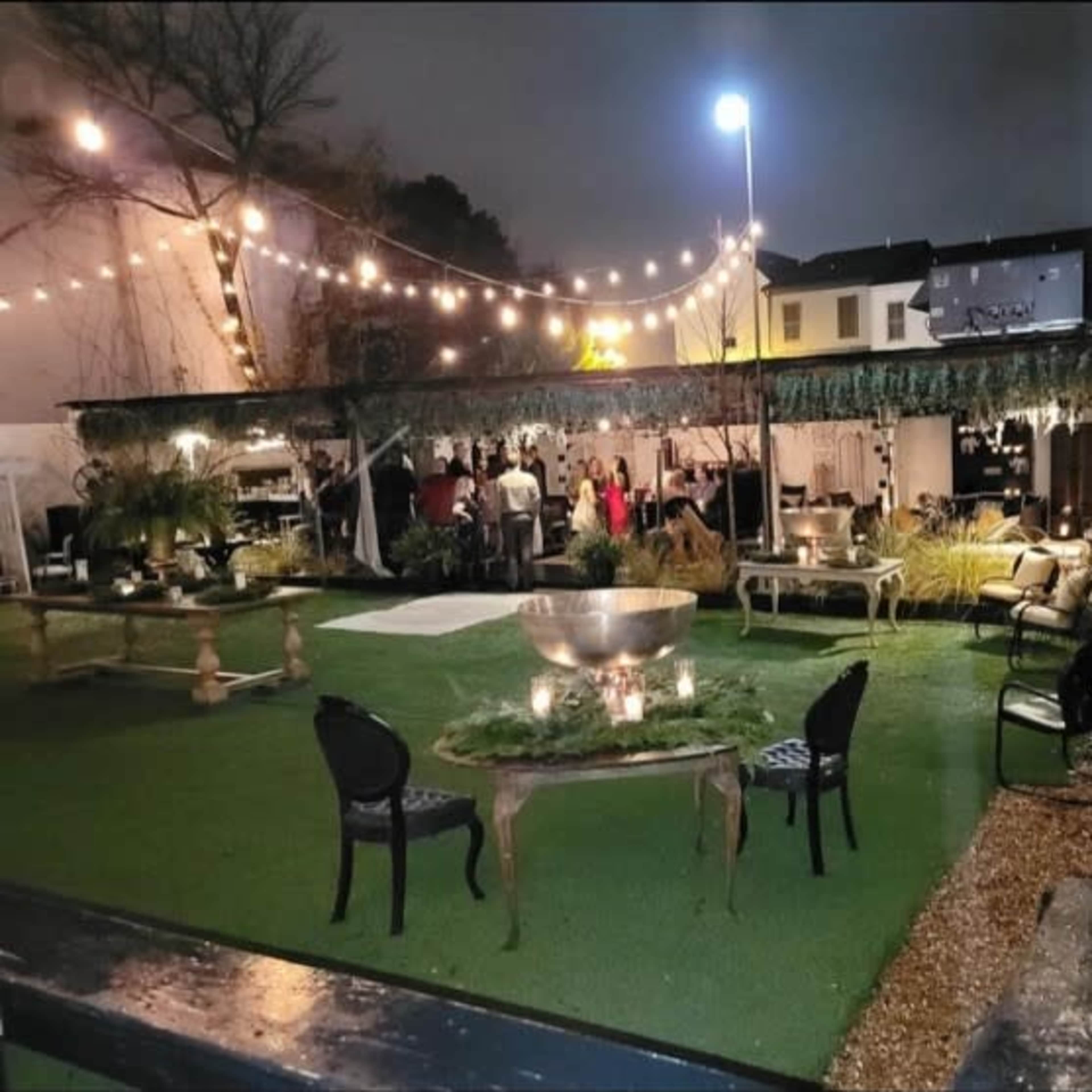 An outdoor event space with string lights, tables, and seating arranged on a lawn, creating a festive atmosphere.