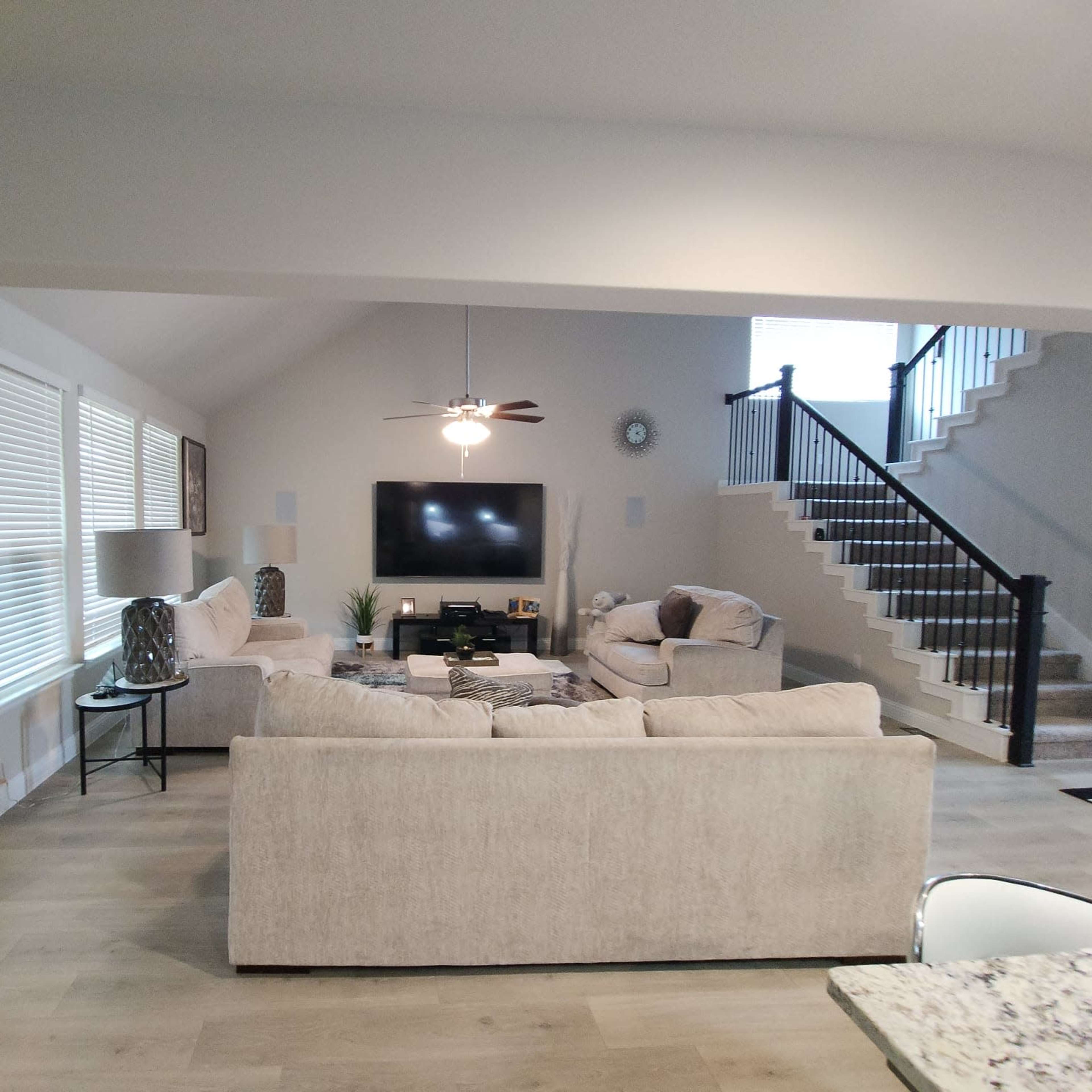 A spacious living room with two light-colored sofas facing a wall-mounted television, adjacent to a staircase leading to an upper level.