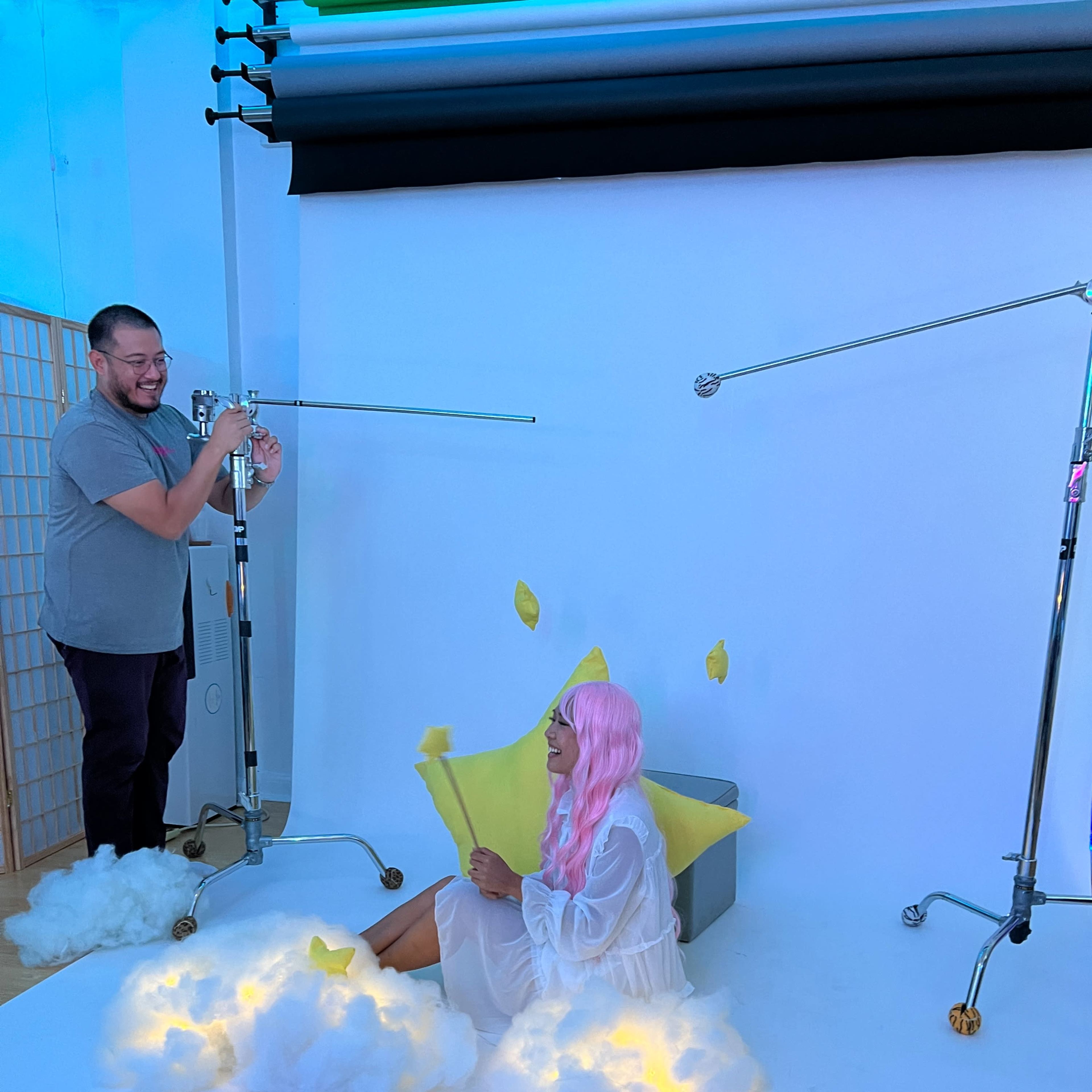 A person with pink hair sits on a cloud-like prop while another individual adjusts lighting equipment in a photography studio setting.