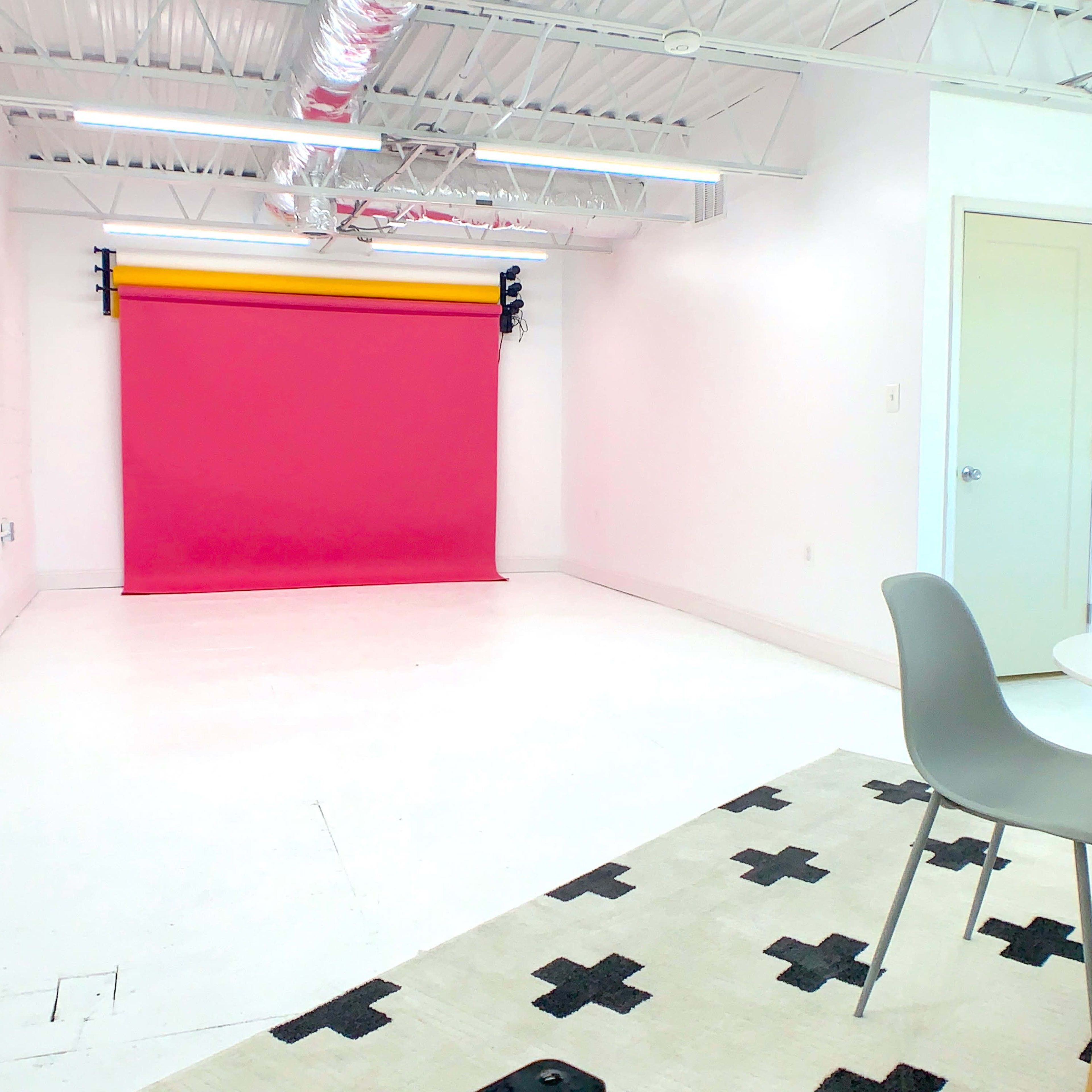 A bright, empty studio space featuring a pink backdrop, white walls, and a small table with chairs on a patterned rug.