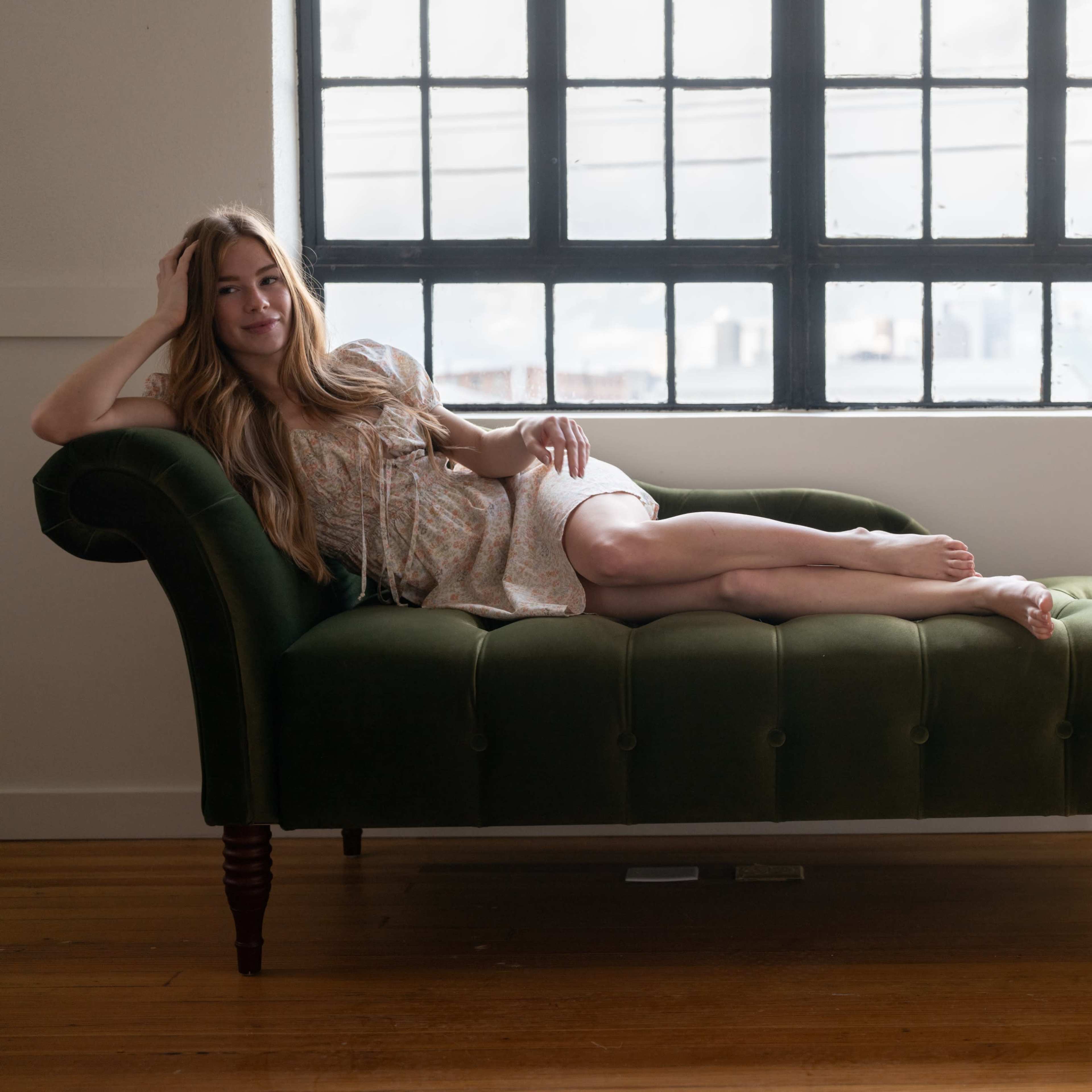 A woman with long hair is reclining on a green chaise lounge next to a large window.