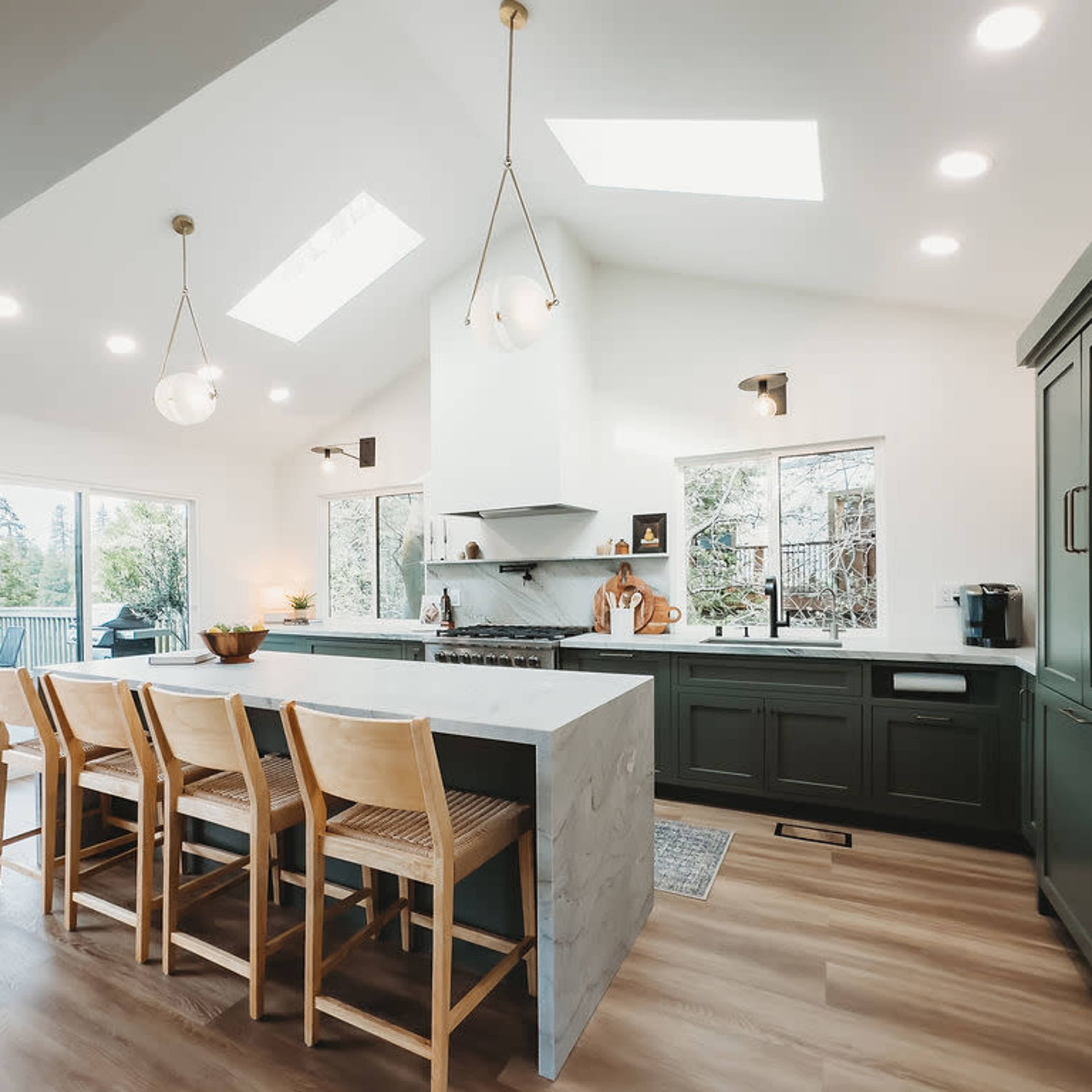 A modern kitchen features a central island with seating, large windows for natural light, and minimalist cabinetry in green and white.