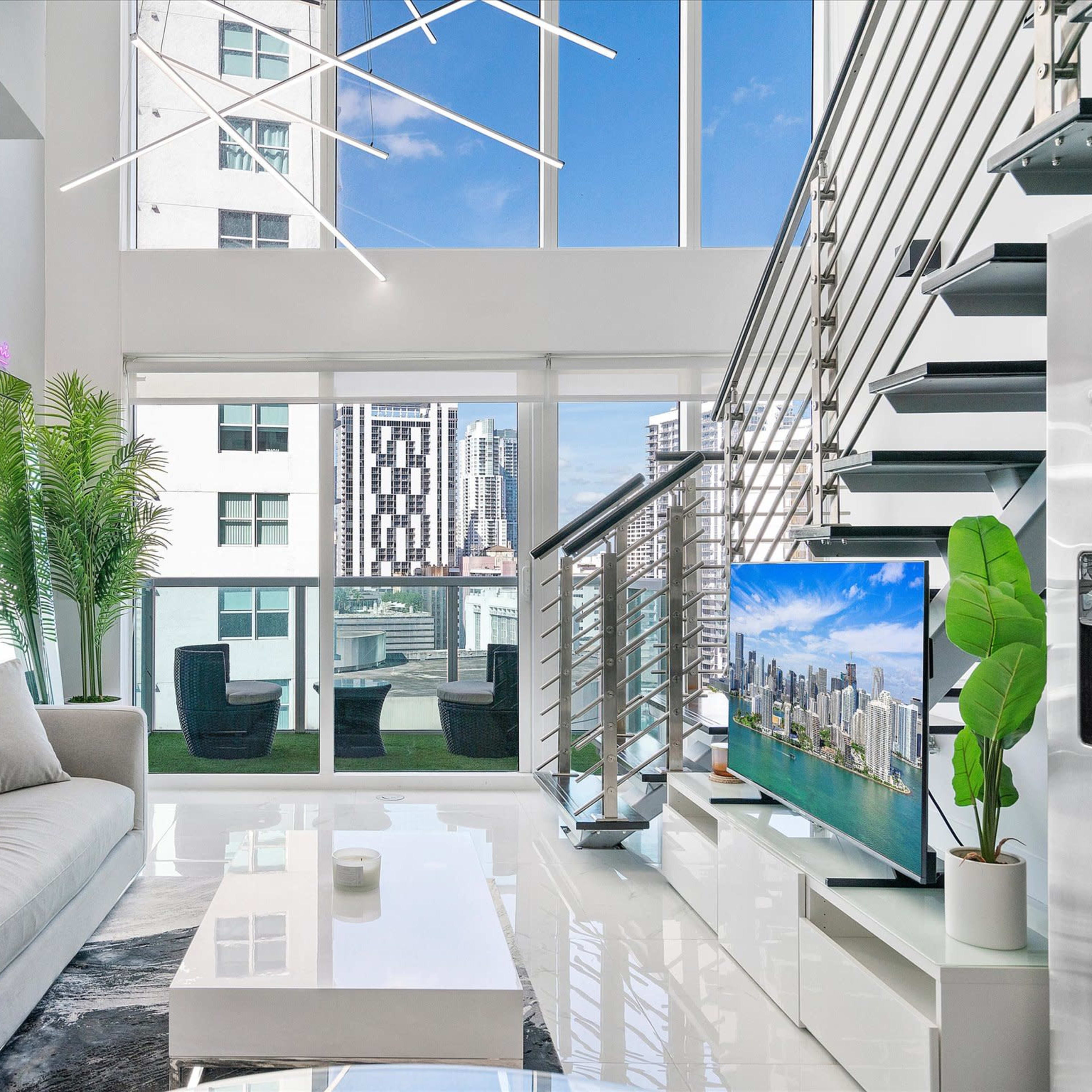 A modern living room with large windows, a sleek gray sofa, a glass coffee table, and a staircase leading to an upper level, overlooking a city skyline.