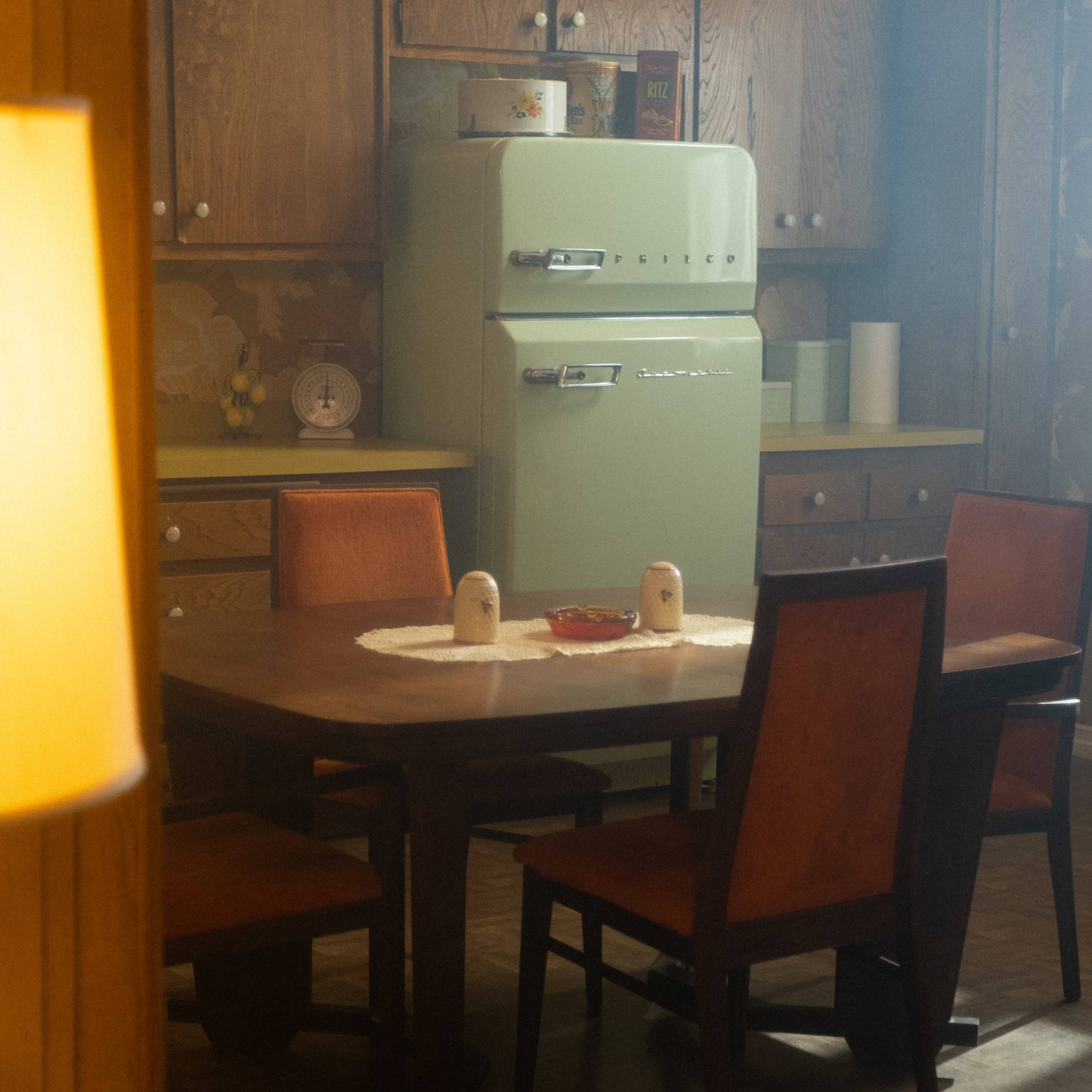 A retro kitchen features a green refrigerator, a wooden dining table with four chairs, and a floral-patterned wallpaper.