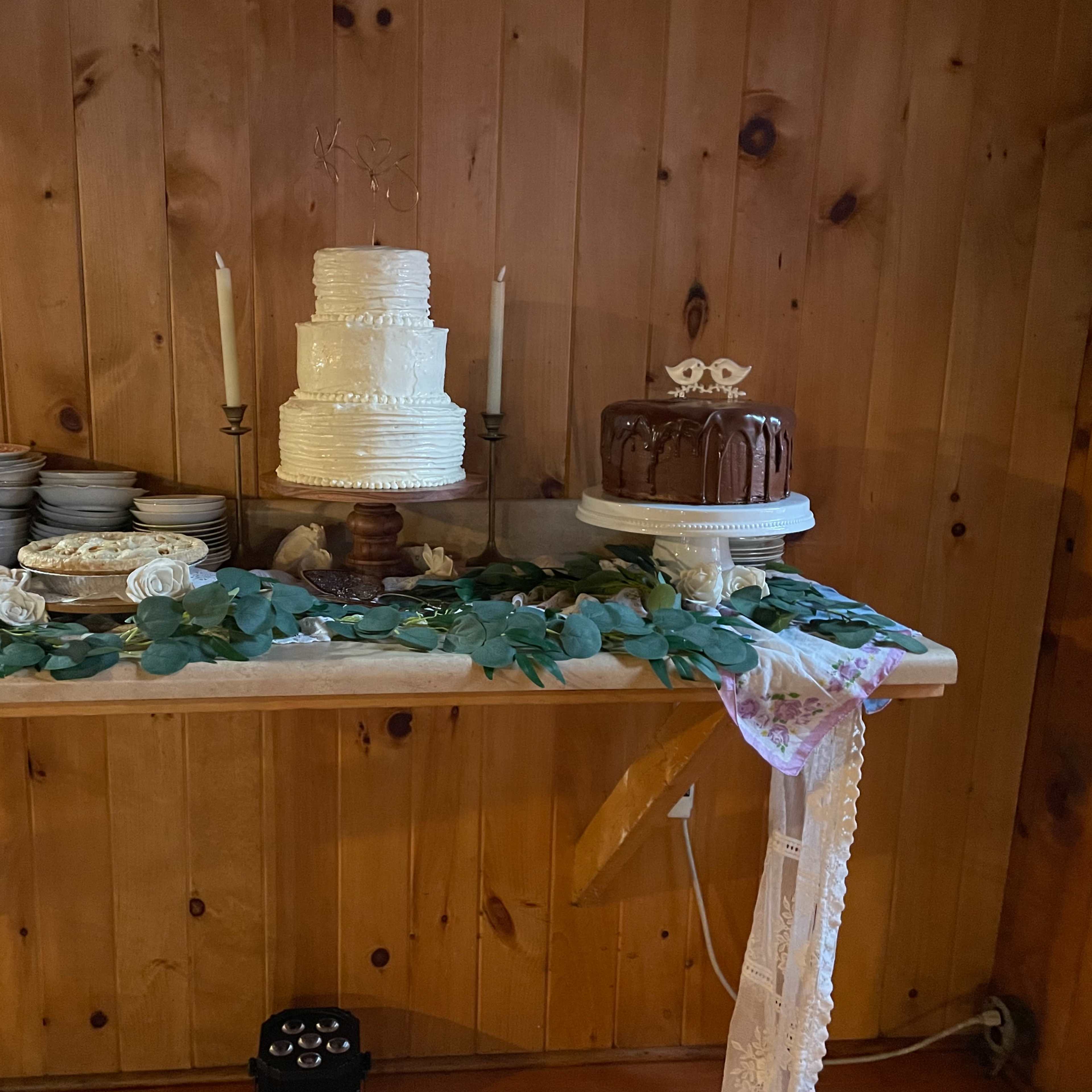 A wooden table displays two wedding cakes, a white tiered cake and a chocolate cake, surrounded by plates and decorative greenery.