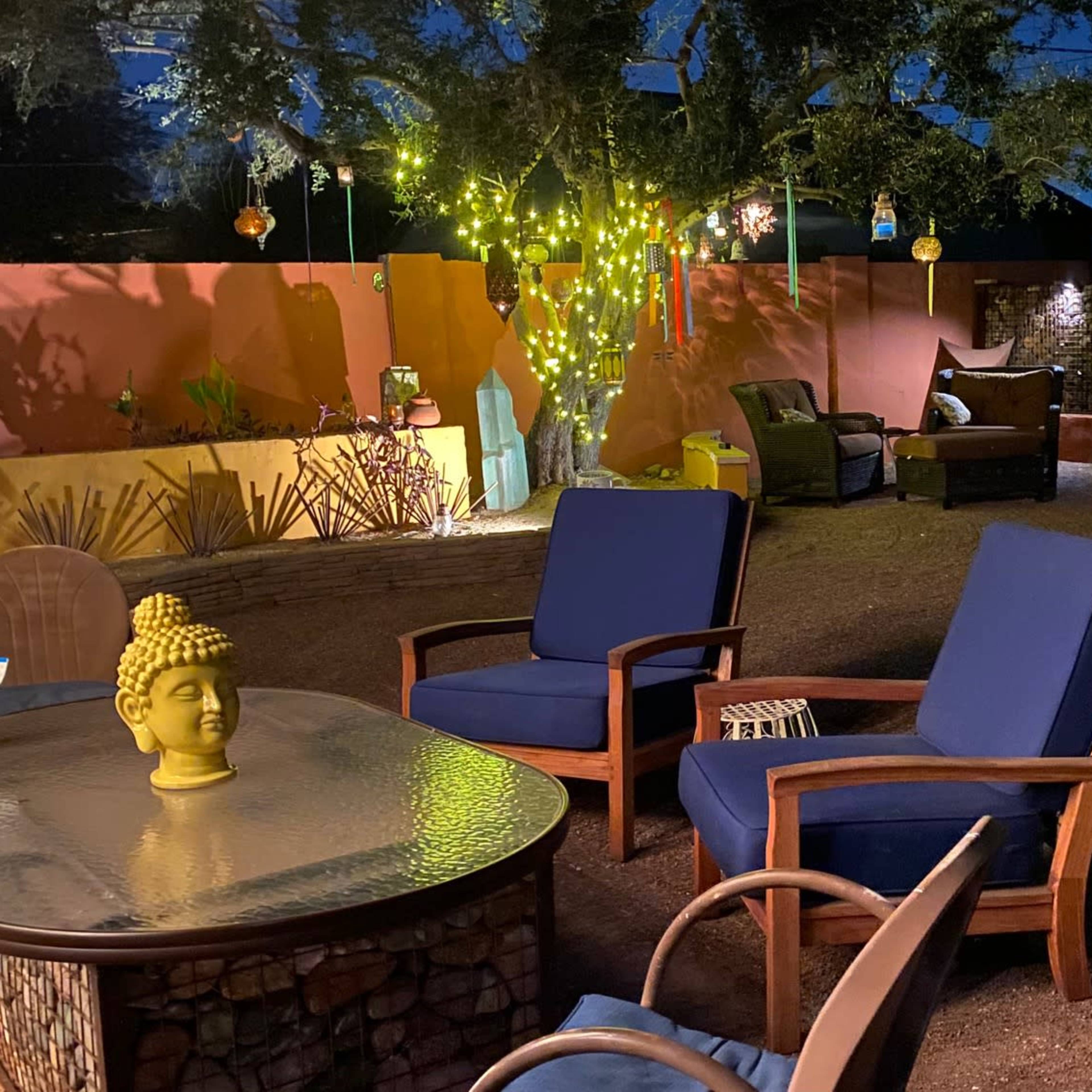 A landscaped outdoor patio at night, featuring a table with a Buddha statue and two blue chairs, surrounded by decorative lights and plants.